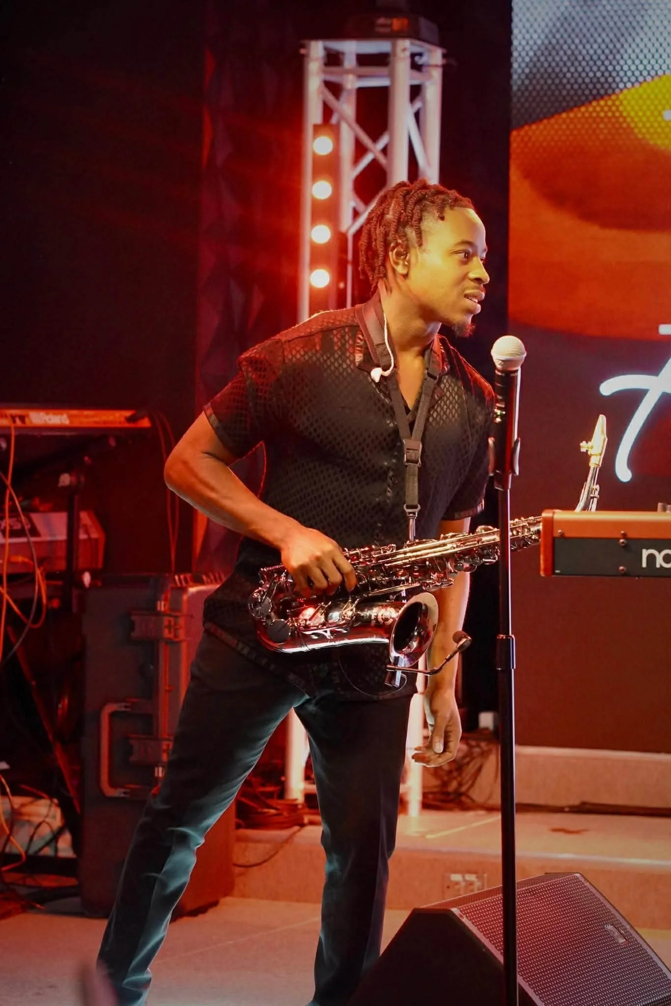 A man with dreadlocks holding a microphone and a saxophone on stage, with a stage rig and lighting in the background.