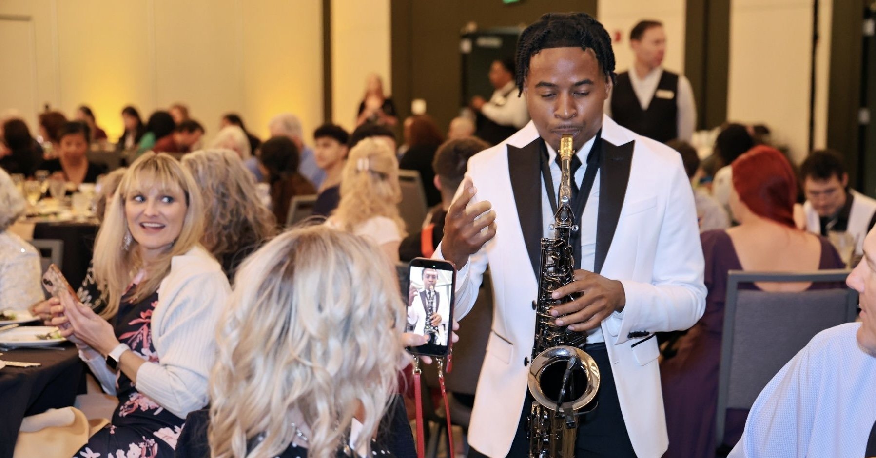 A man in a white tuxedo jacket playing the saxophone at a formal event with many guests seated at tables in the background.