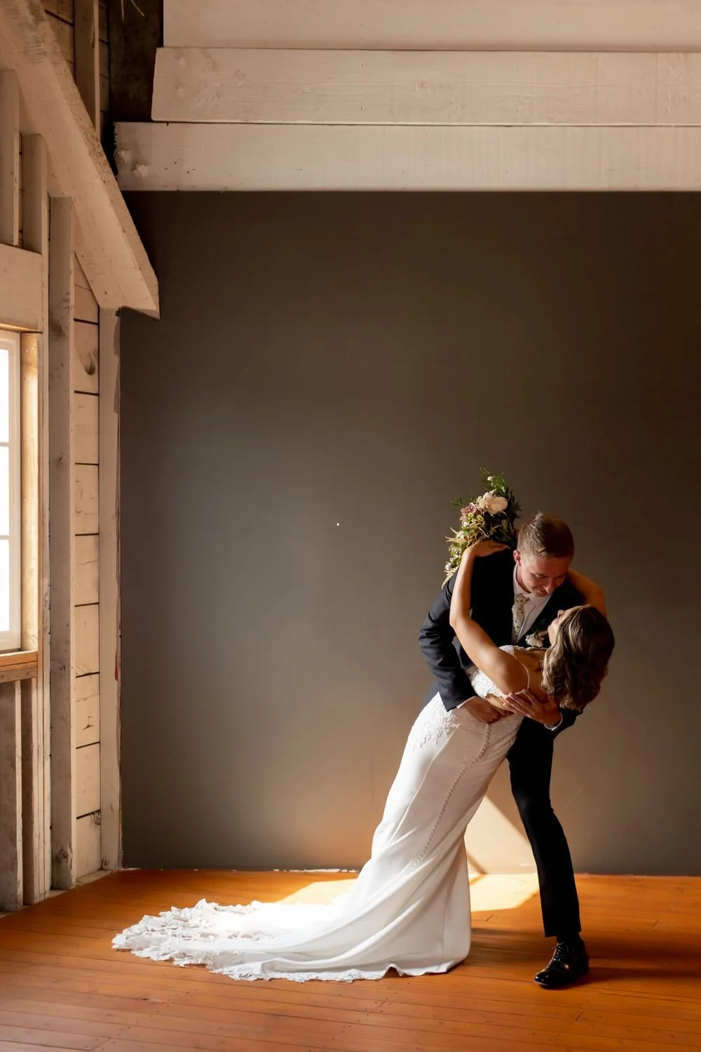 A couple dancing at their wedding, with the groom dipping the bride while holding her waist, in a rustic wooden venue, both smiling.