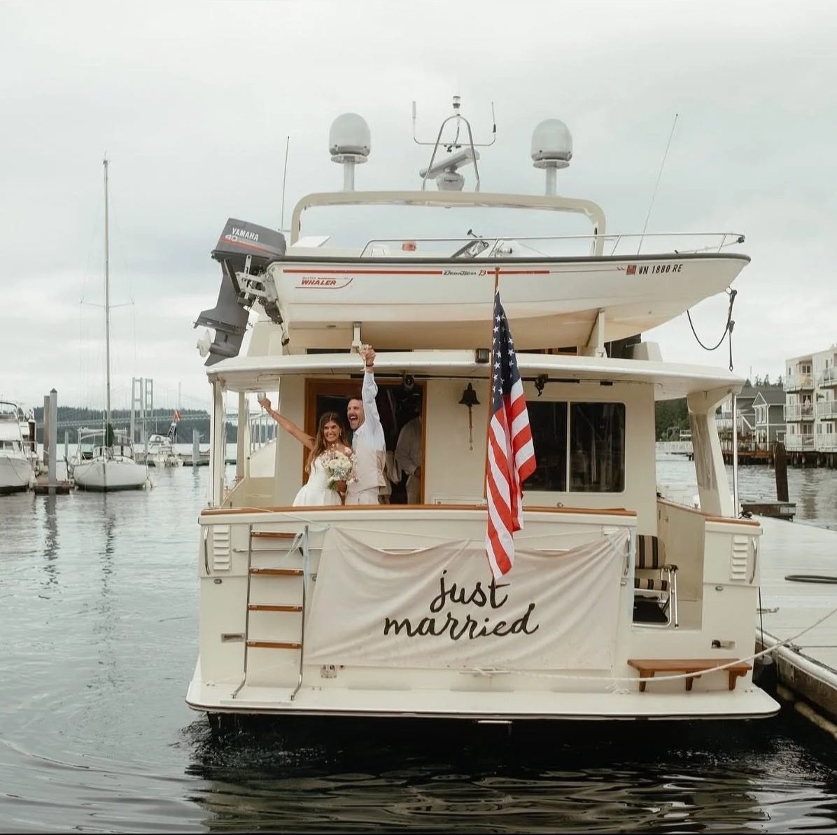 A newlywed couple celebrating on a boat with a 'Just Married' banner, American flag, and marina background.