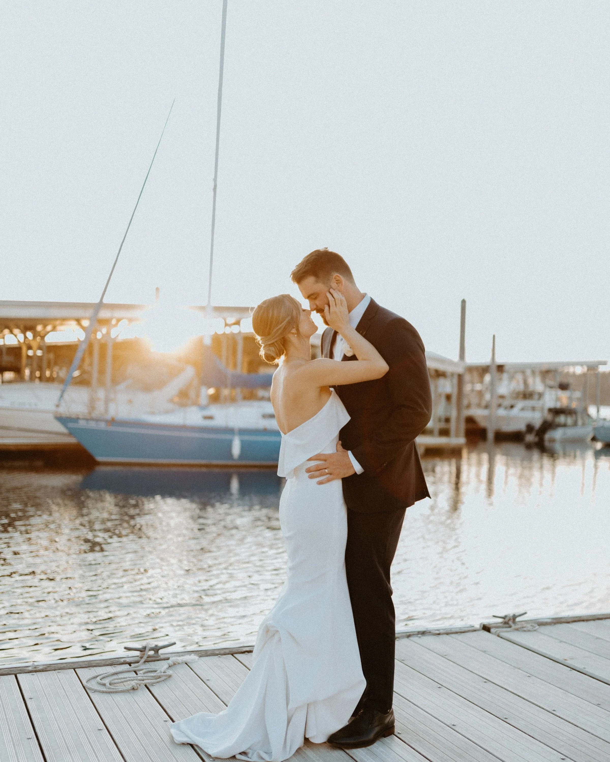 A bride and groom share a dance on a dock by the water during sunset, with boats and a marina in the background.