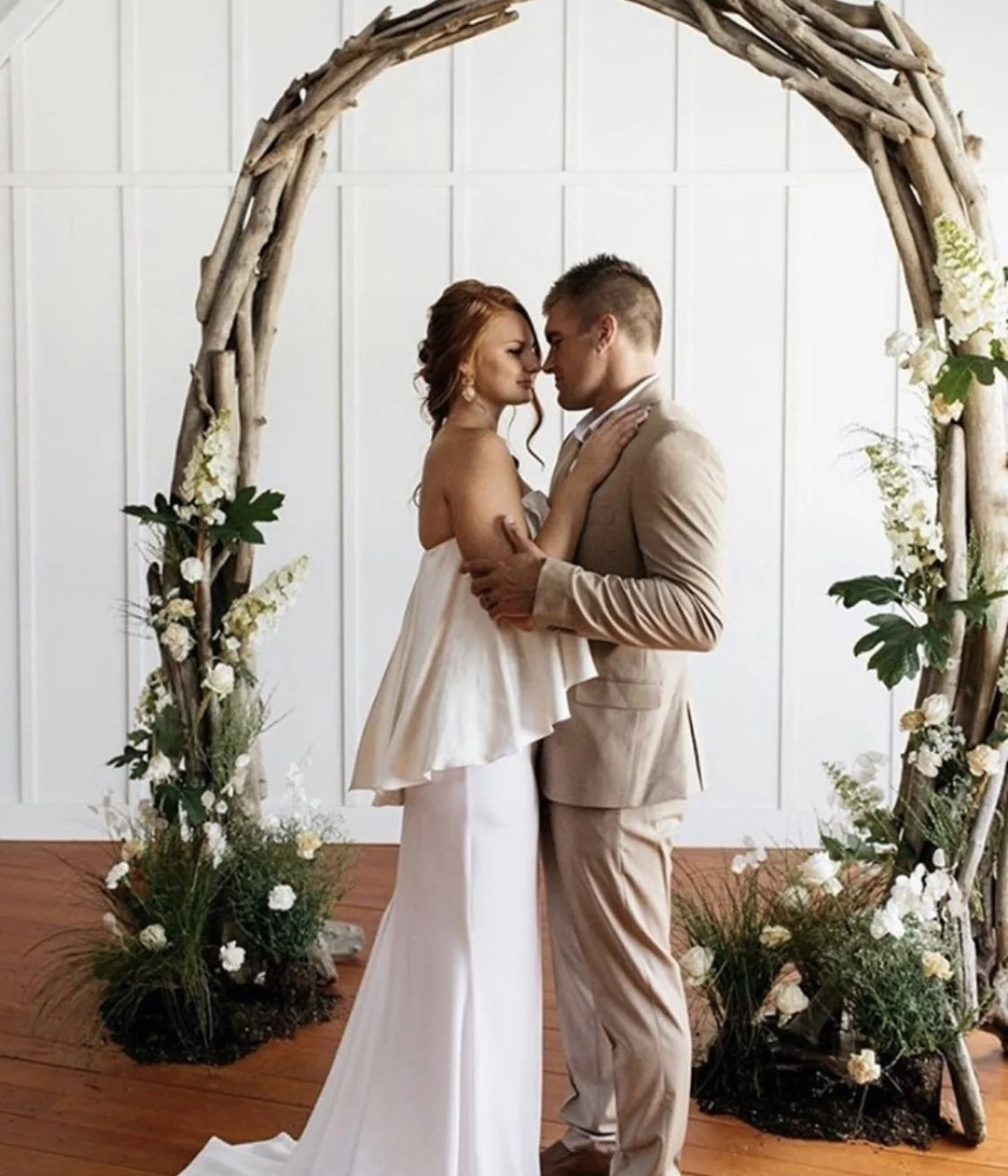 A bride and groom embrace under a wooden floral arch at their wedding ceremony, with white flowers and greenery decorating the arch.