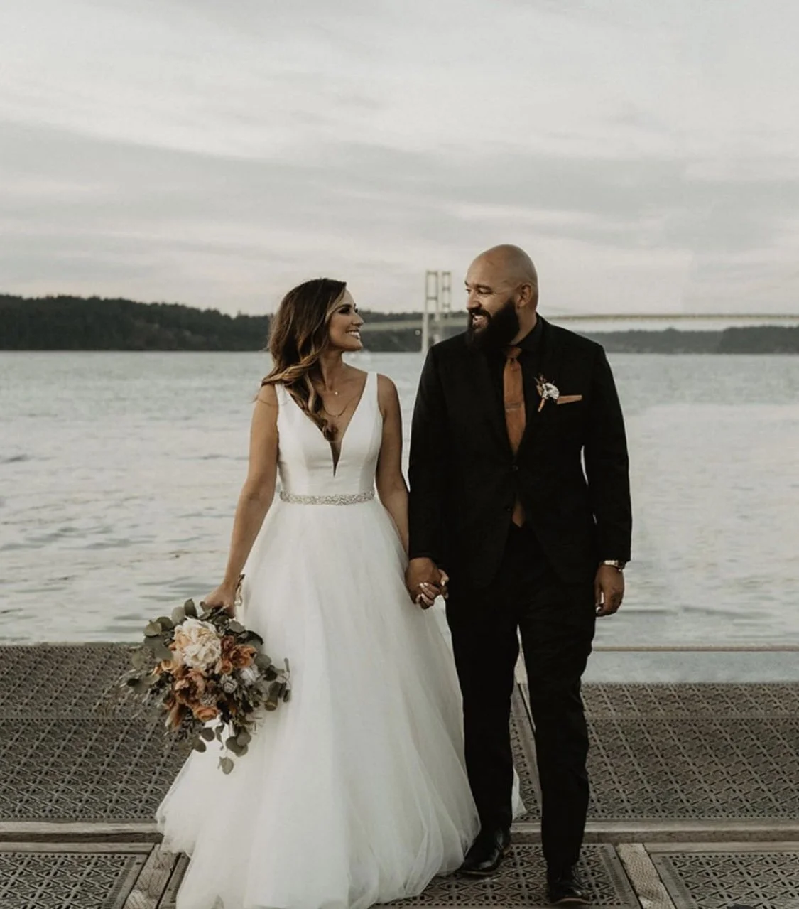 A newlywed couple holding hands and smiling at each other by the water, with a bridge in the background.