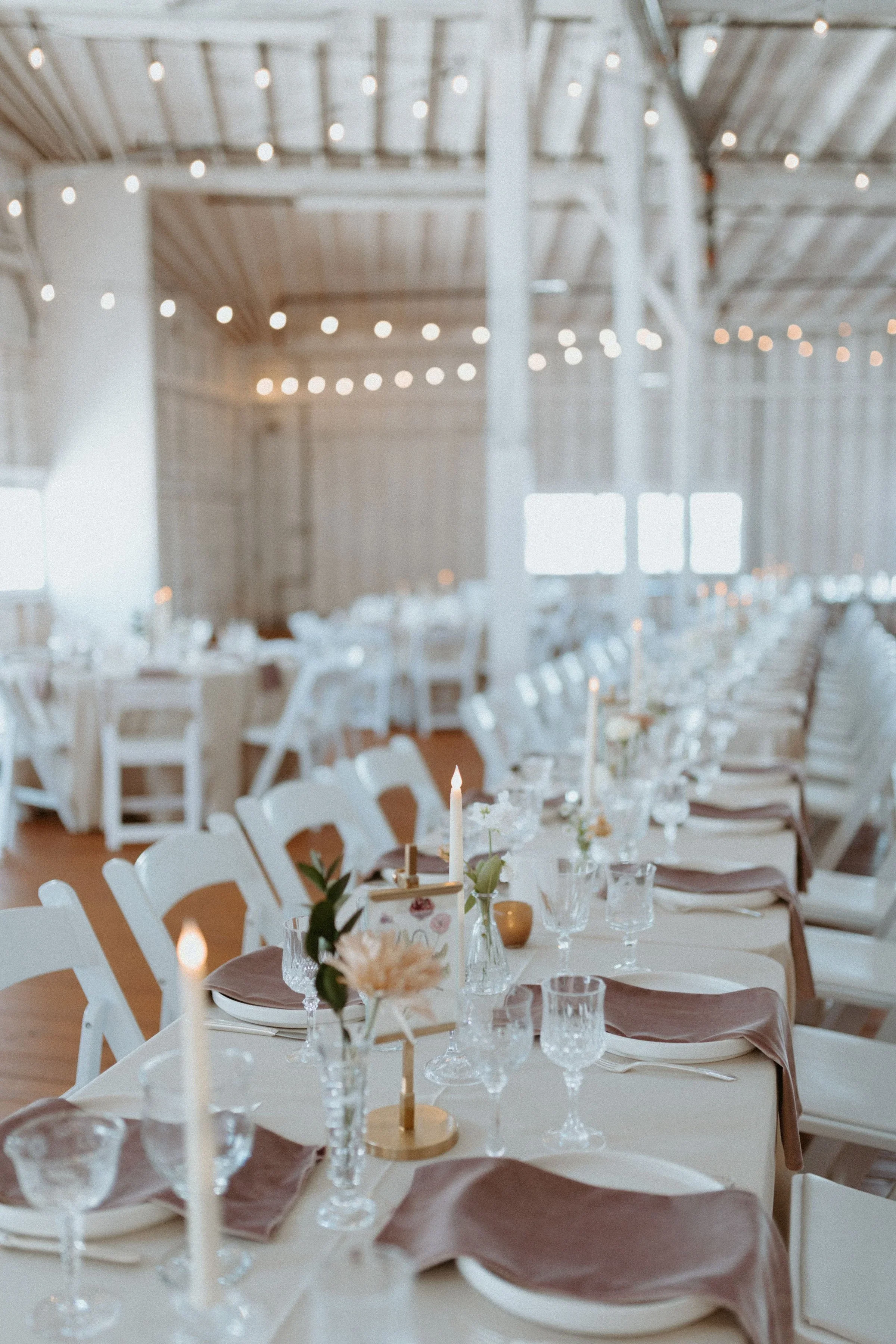 A decorated event space with tables set for a formal occasion, featuring floral centerpieces, candles, and glassware, with string lights hanging from the ceiling.