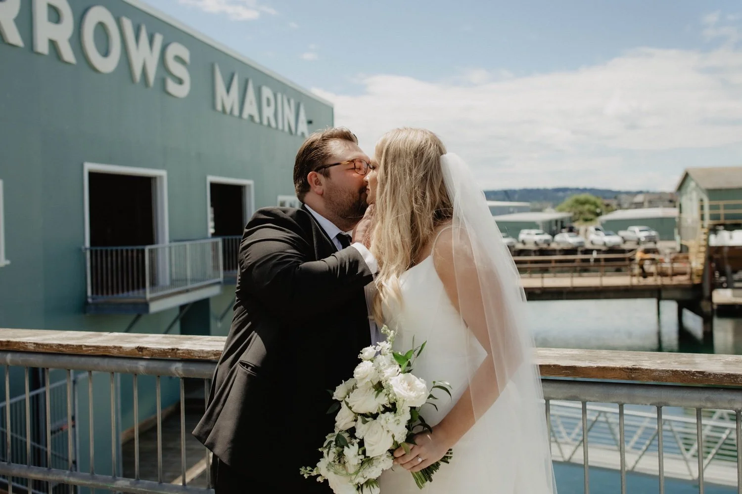 A bride and groom sharing a kiss outdoors near a marina with parked cars and buildings in the background.