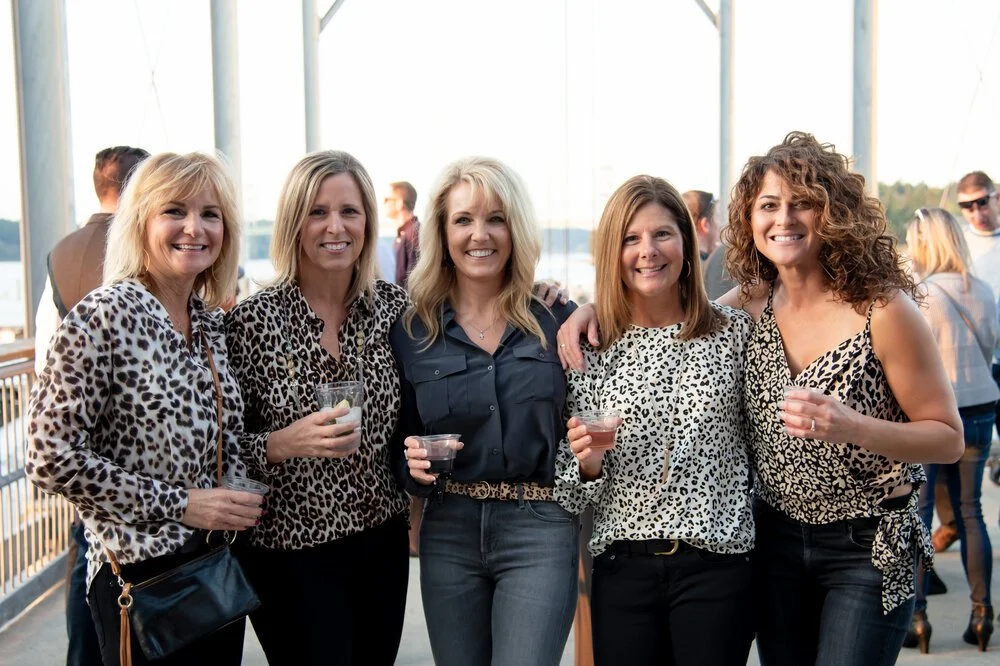 Five women standing together celebrating on a boat deck, holding drinks, wearing animal print clothing.
