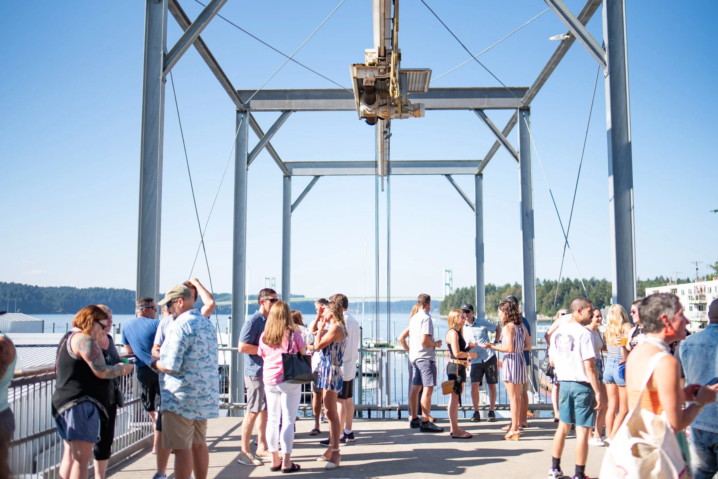 Group of people standing on a dock near water, with a metal framework overhead and a bridge in the background, on a sunny day.
