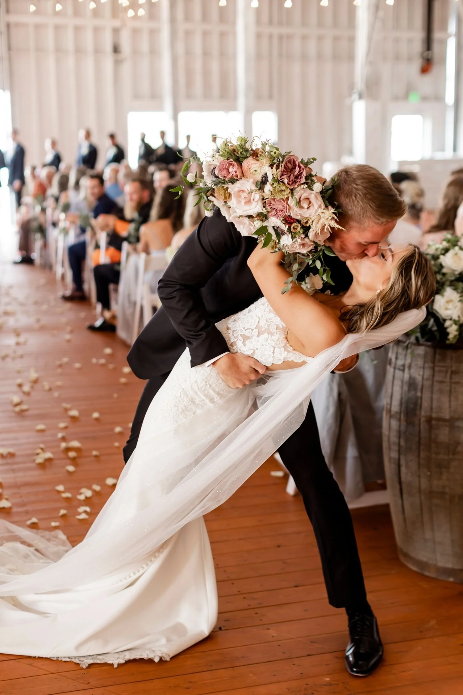 A bride and groom share a kiss during their wedding ceremony in a rustic barn with wooden floors and walls. The groom is dressed in a black suit, and the bride is wearing a white wedding dress and veil. The groom is bending down while holding the bride, and the bride has her hand on the groom's neck. The bride has a large bouquet of pink, white, and green flowers. Guests are seated in the background, with some groomsmen lined up near the window. Rose petals are scattered on the floor.