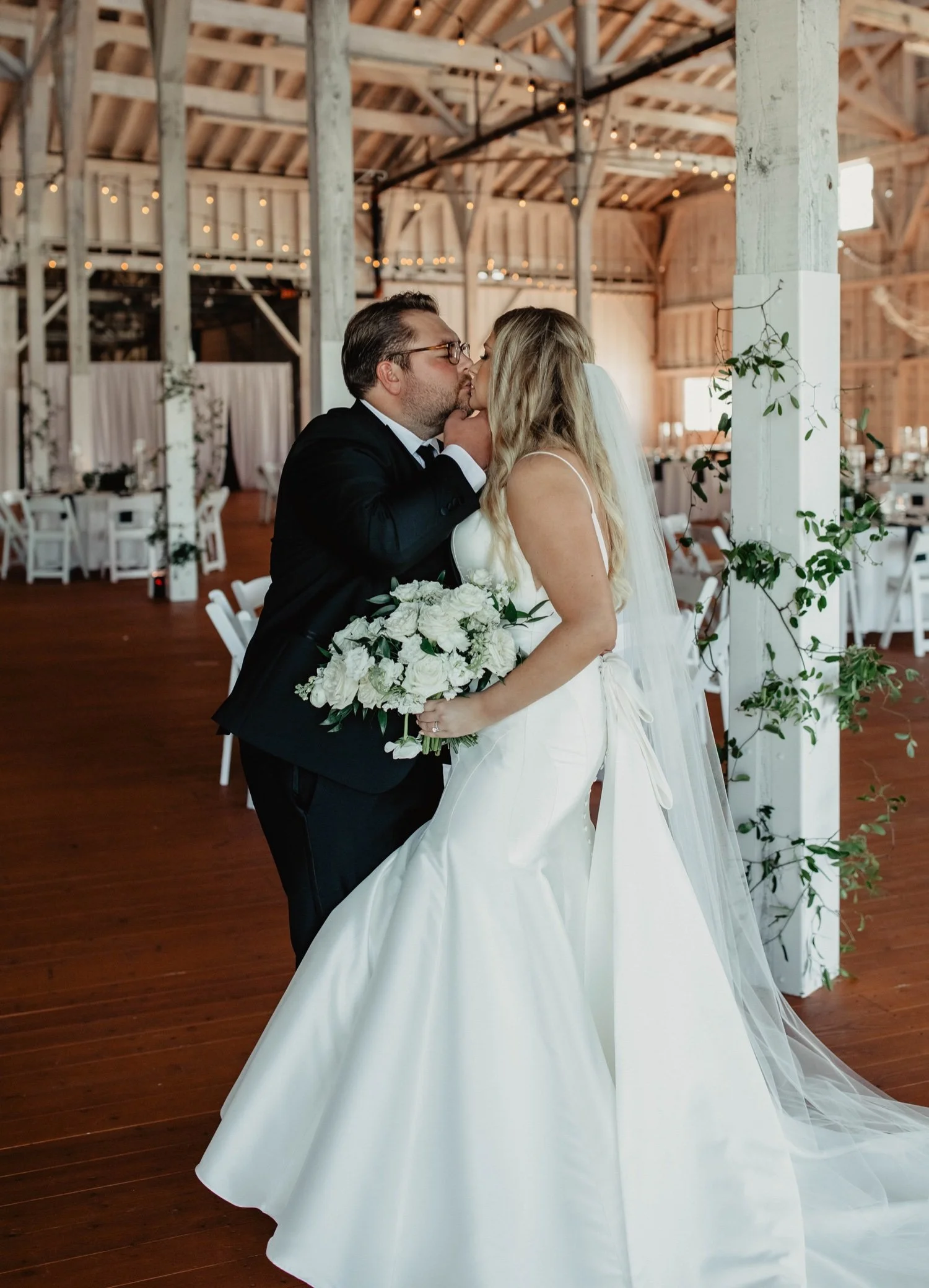 A bride and groom sharing a kiss during their wedding ceremony inside a rustic barn decorated with string lights and greenery.