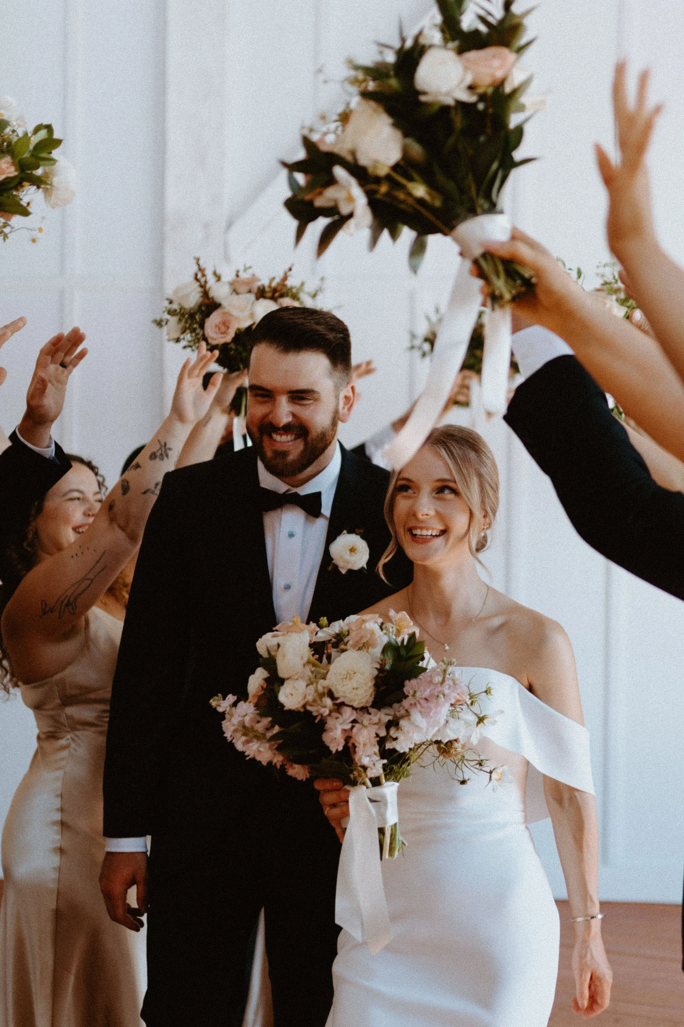 A wedding party celebrating with a bride and groom in formal attire, surrounded by friends holding bouquets and raising their arms in celebration.