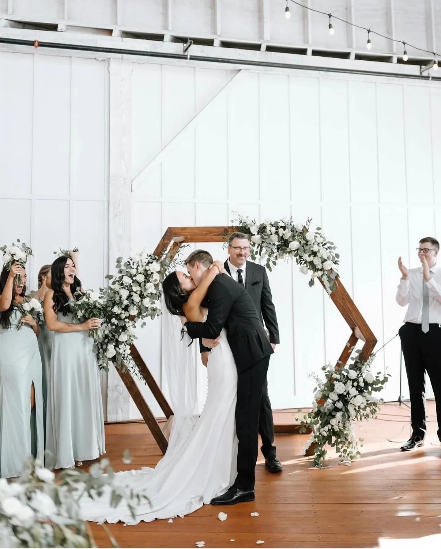 A bride and groom kiss during their wedding ceremony as officiant looks on, surrounded by bridesmaids holding bouquets, with decorative floral arrangements and a geometric wooden arch in the background.