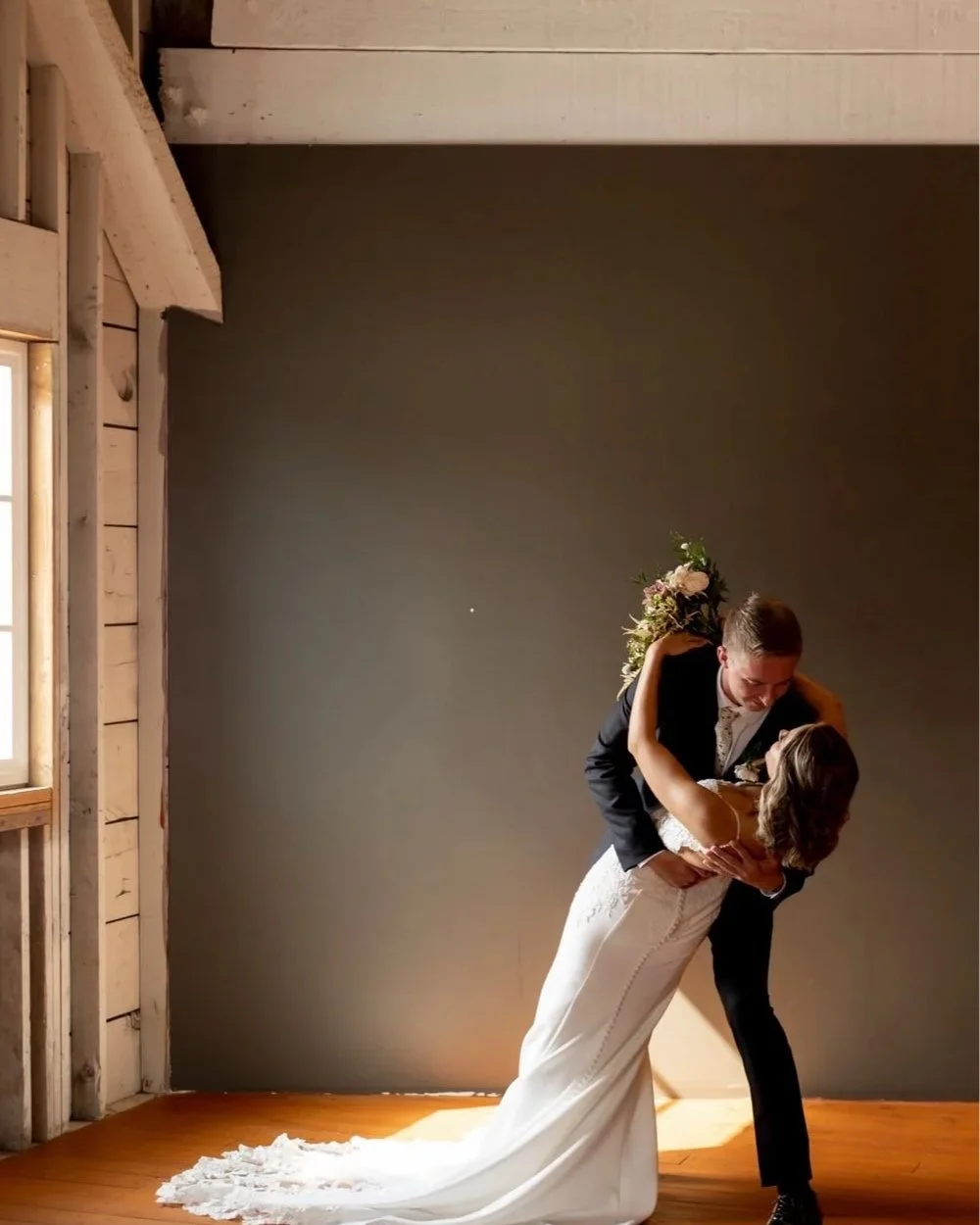 A newlywed couple dancing indoors, with the groom dipping the bride. The bride is wearing a white wedding dress with a train, and the groom is in a black suit. She is holding a bouquet behind his neck, and they are embracing happily.