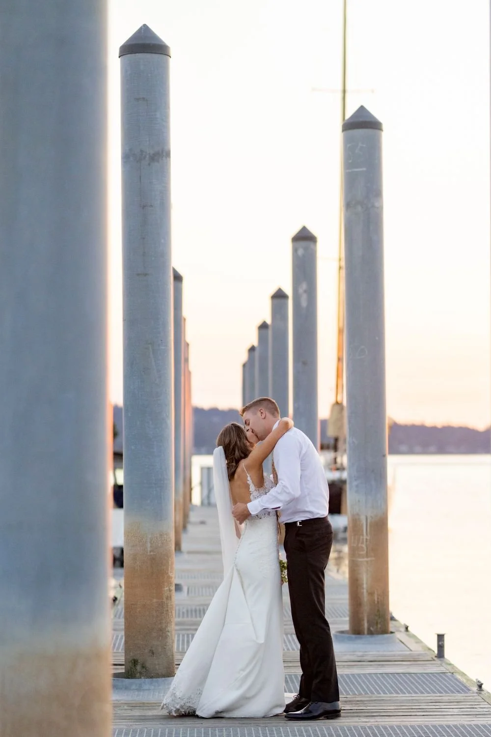 A bride and groom affectionately embrace and kiss on a dock with large metal posts, near a body of water during sunset.