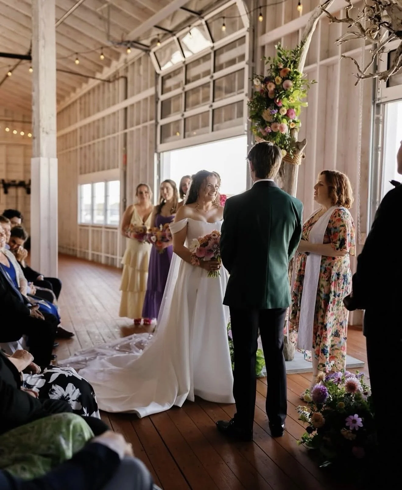A wedding ceremony taking place in a rustic wooden venue with sunlight streaming through large windows. A bride and groom are exchanging vows, with the bride holding a bouquet of pink and white flowers. Bridesmaids in colorful dresses are standing behind them, holding flowers. Guests are seated on both sides, watching the ceremony. There are floral arrangements and hanging lights decorating the space.