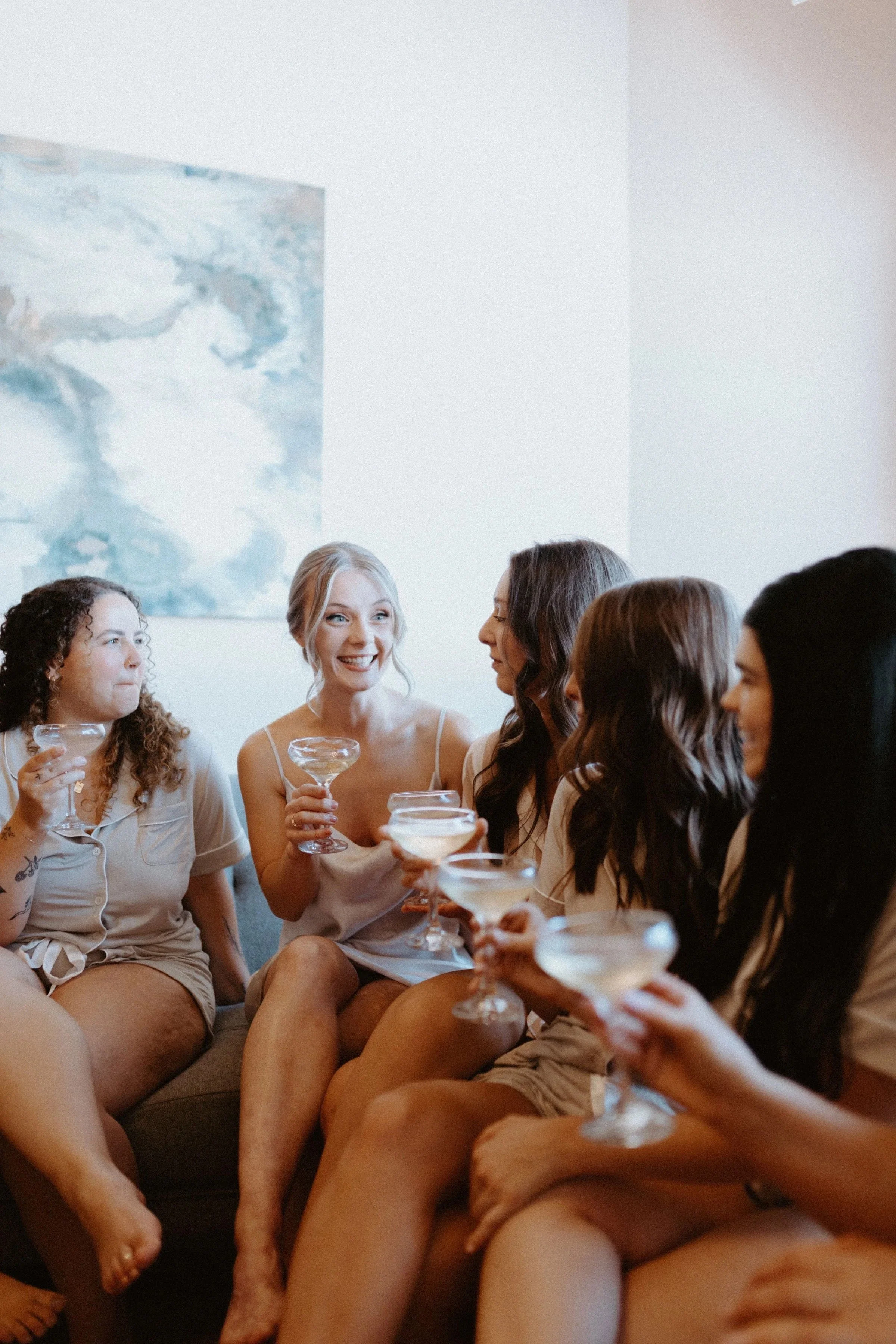 A group of women sitting on a sofa and chairs, wearing casual sleepwear, holding cocktails, and enjoying a social gathering in a room with a painting on the wall.