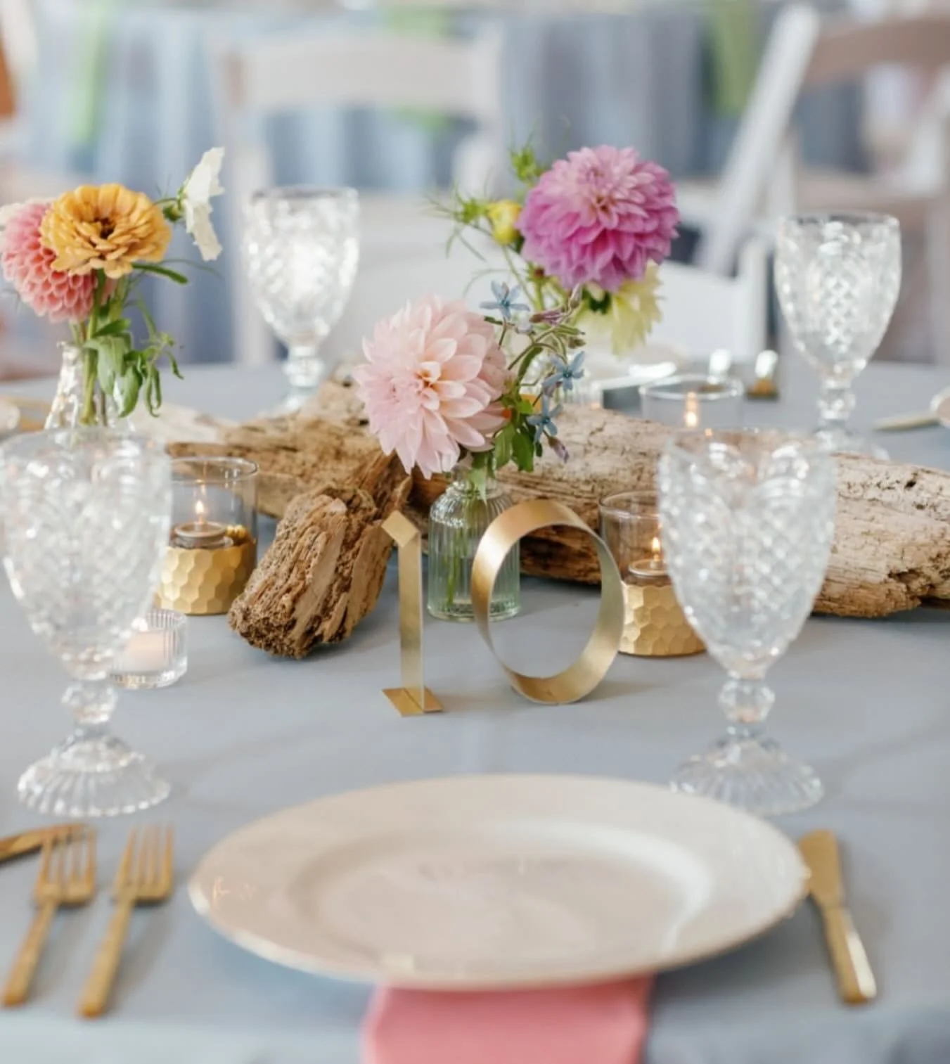 A table decorated for a wedding or celebration with pink and yellow flowers in a clear glass vase, surrounded by gold-patterned candles, clear engraved glass goblets, and a gold number "10" centerpiece on a light gray tablecloth.