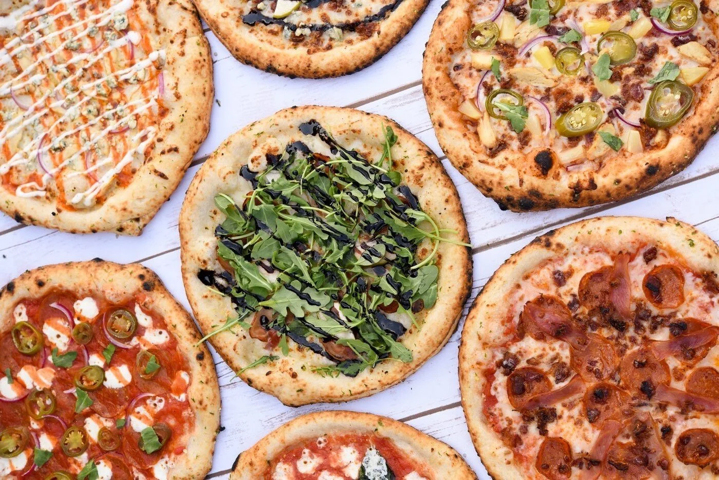 Several small pizzas with various toppings on a white wooden surface.