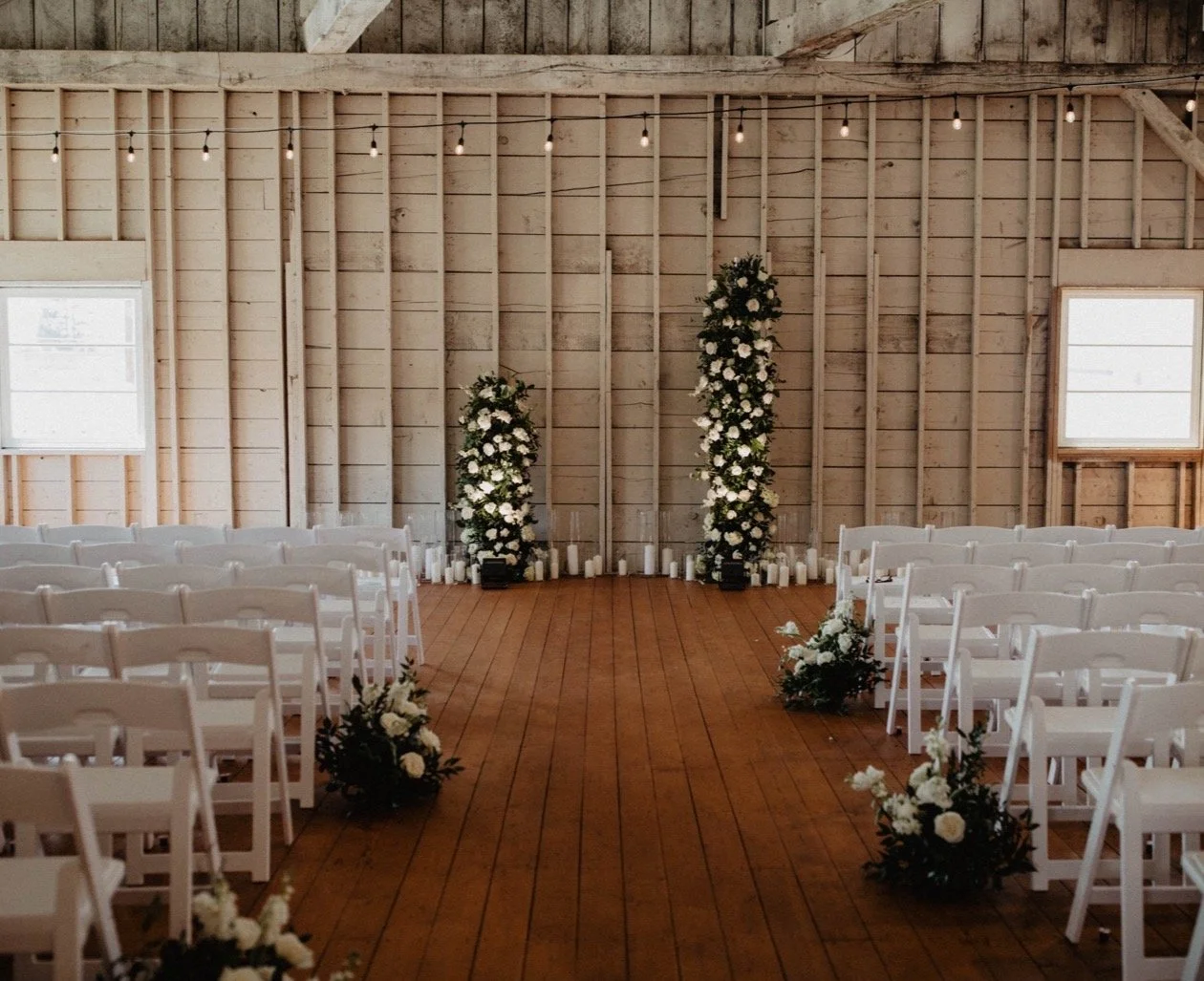 Indoor wedding ceremony setup with white chairs, floral arrangements, and candle decorations on a wooden floor with a rustic wooden backdrop.