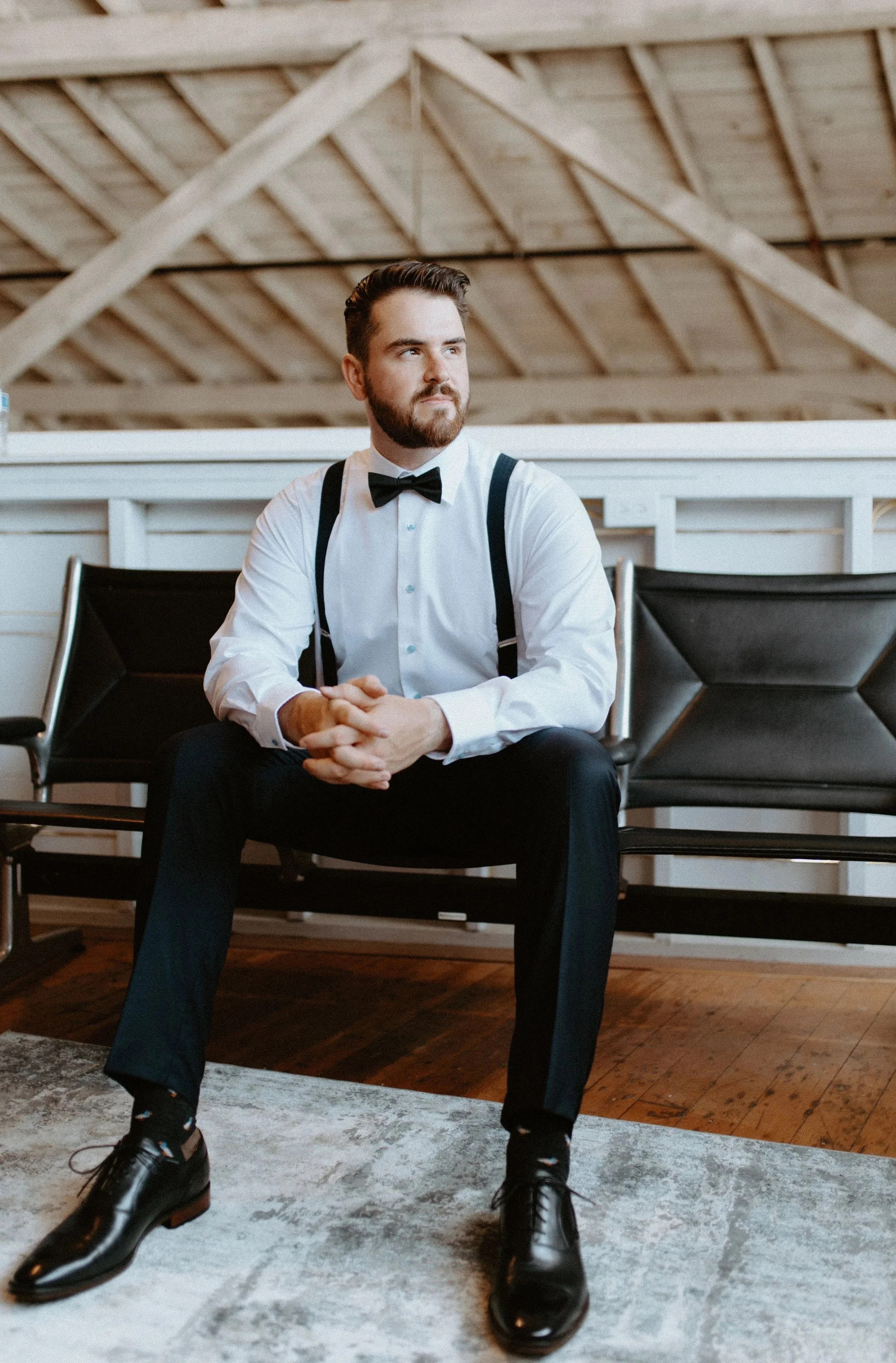 Young man in formal attire sitting on a black chair in a room with a wooden ceiling, wearing a white dress shirt, black suspenders, black bow tie, black pants, and shiny black shoes.