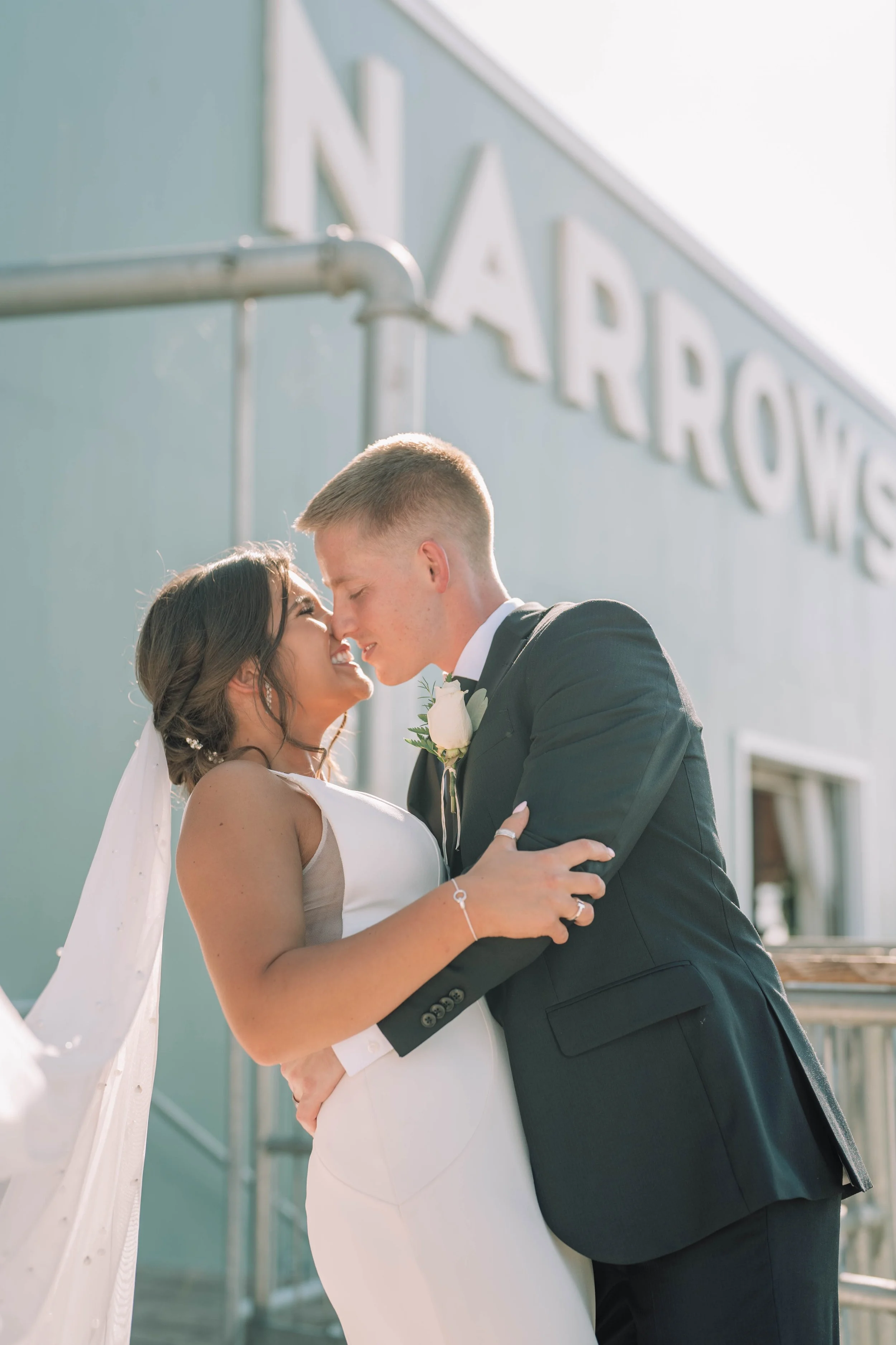 A bride and groom in wedding attire standing close together, smiling and touching foreheads outside a building with a sign partially visible in the background.