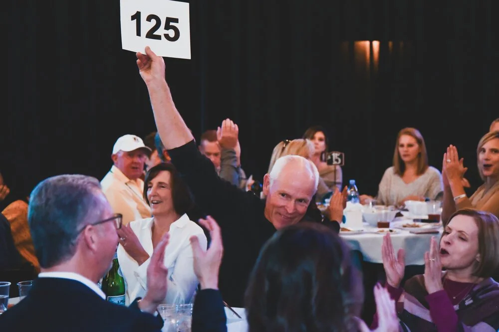 A man with white hair smiling and holding a sign with the number 125 at a banquet table filled with people clapping and celebrating.