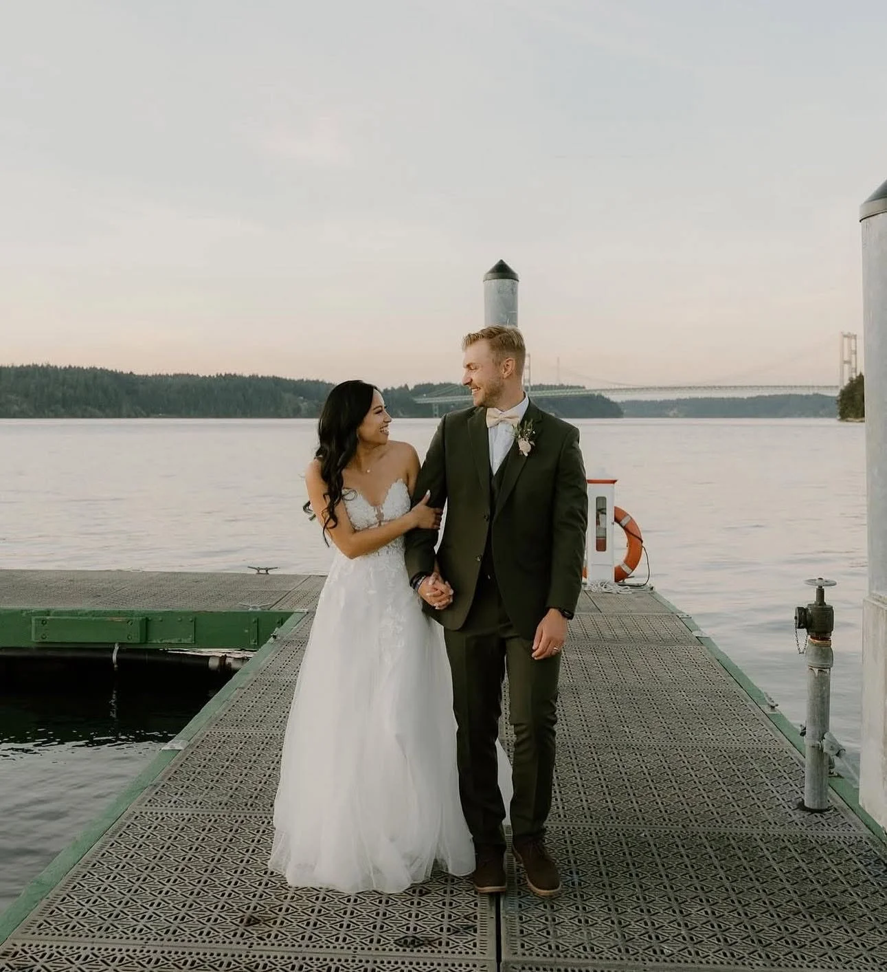 Couple in wedding attire holding hands on a pier by the water, with a bridge in the distance and overcast sky.