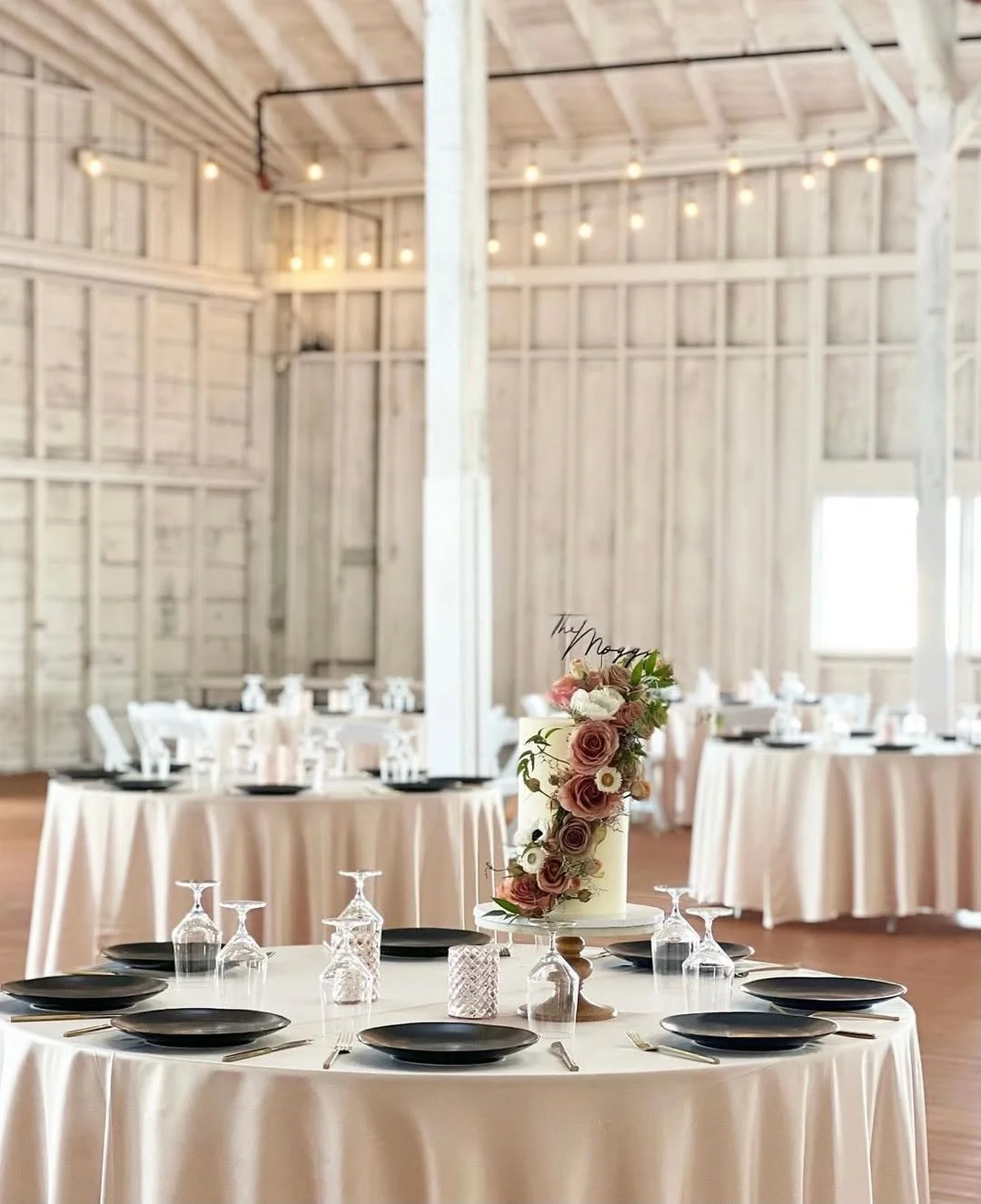 Round table set with black plates, upside-down wine glasses, silverware, a pink and white flower centerpiece, and a 'The Nogy' cake topper in a rustic barn-style venue with string lights and wooden walls.