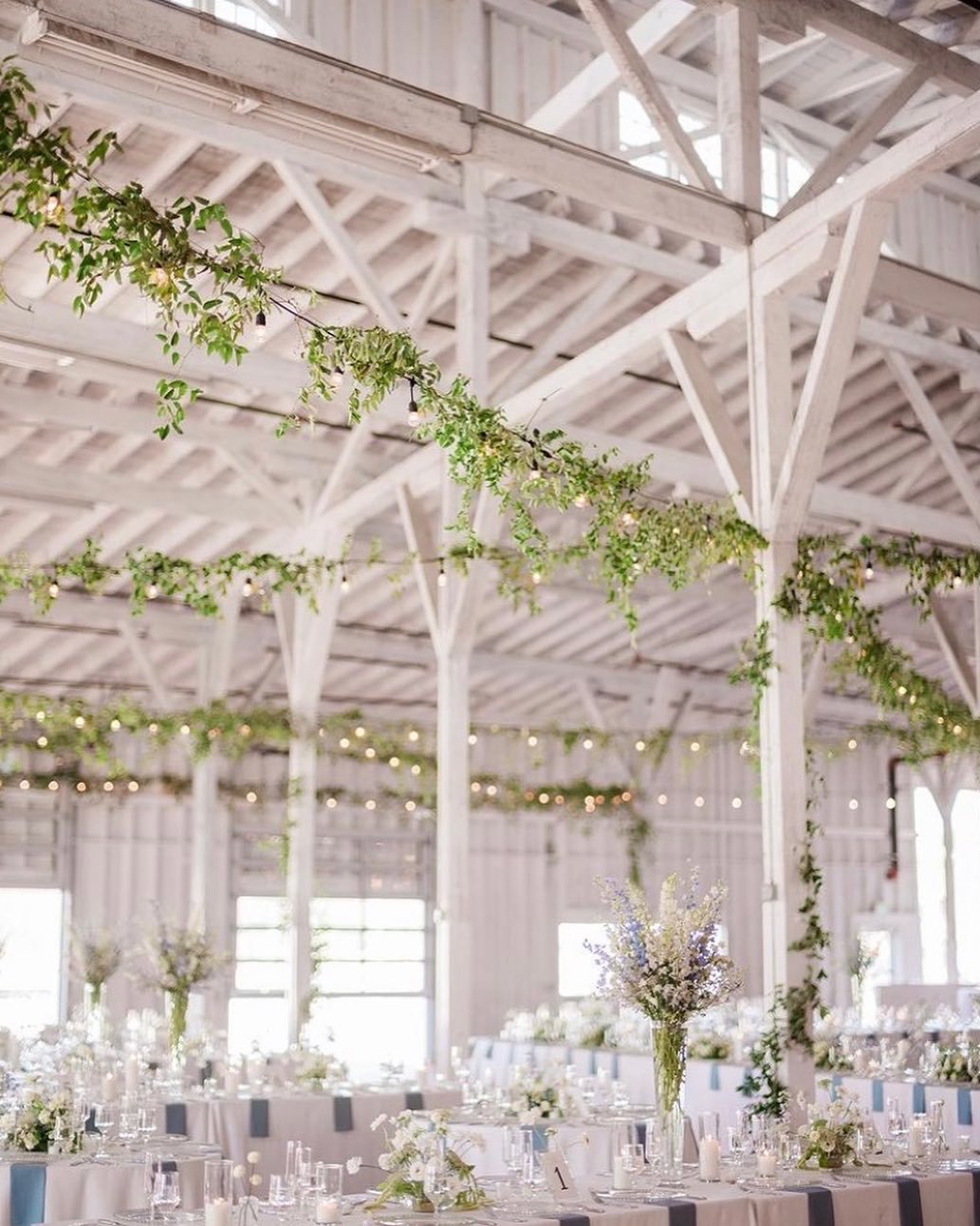 Wedding reception in a bright barn with hanging garlands and string lights, decorated with floral arrangements on tables.