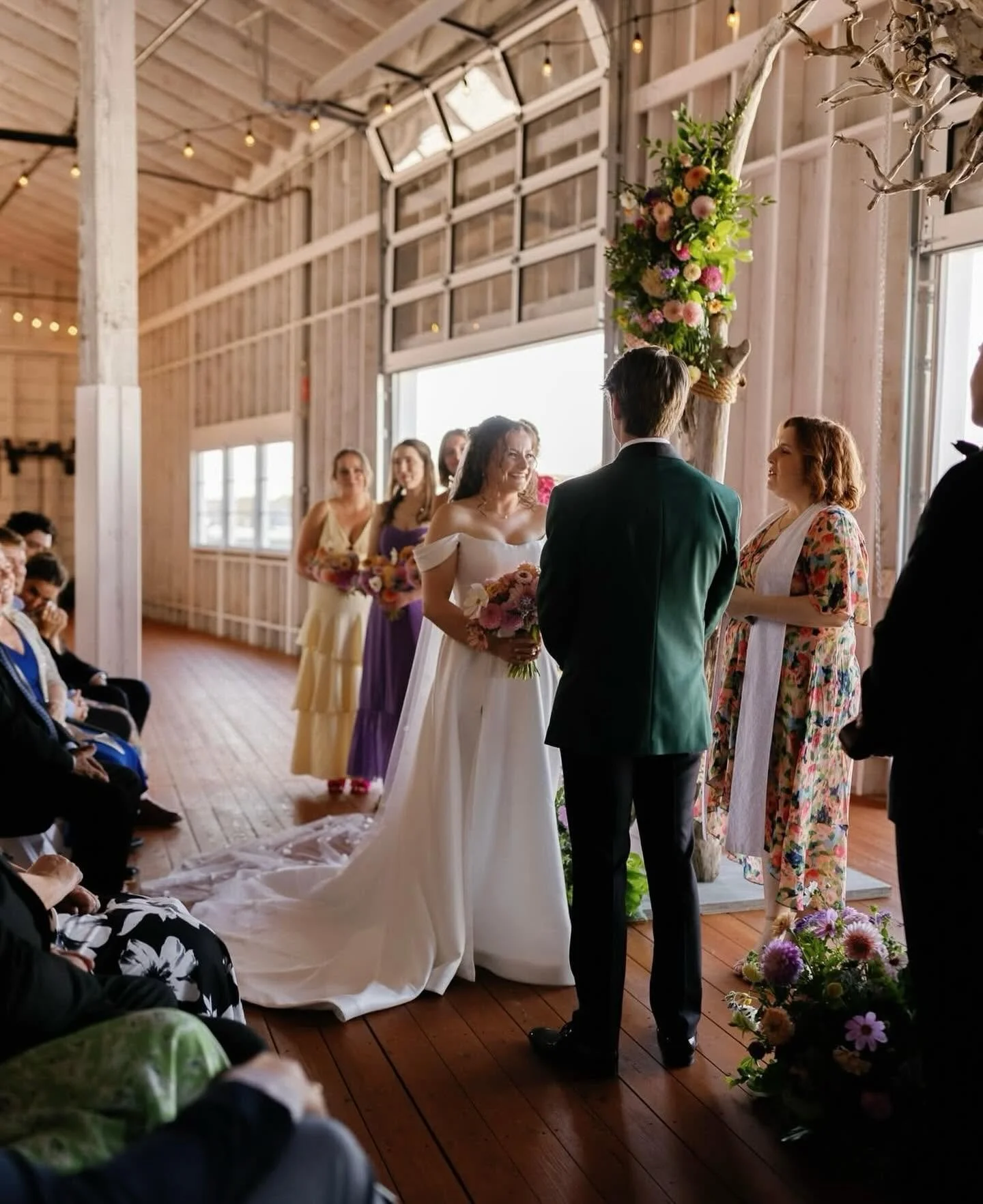 A bride and groom exchange vows during a wedding ceremony in a rustic barn with wood floors and walls, decorated with flowers and string lights.