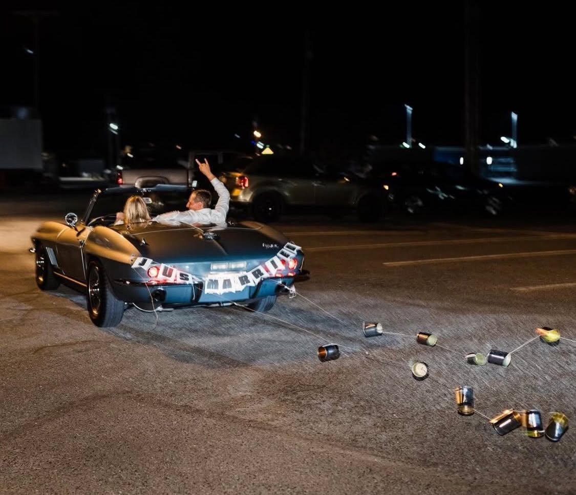 A black convertible car on a street at night with a bride and groom inside, celebrating their wedding with a 'Just Married' sign. Many cans are tied to the back of the car, some are scattered on the ground, and the groom is making a peace sign with his hand.