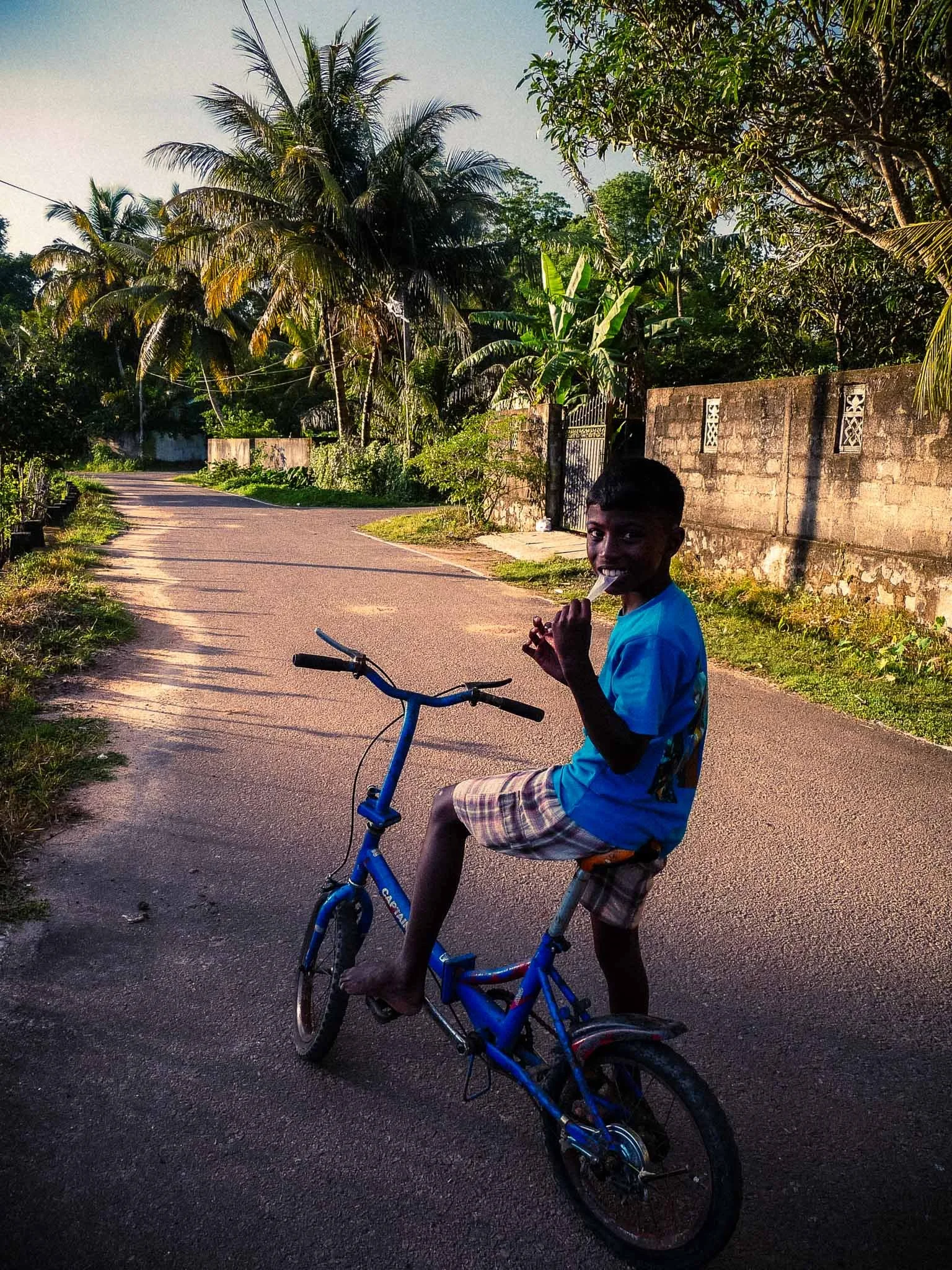 Lanka Surf House — Pitiduwa Village, Weligama — A boy riding a blue bicycle on a paved road surrounded by greenery and palm trees, holding a popsicle and smiling.
