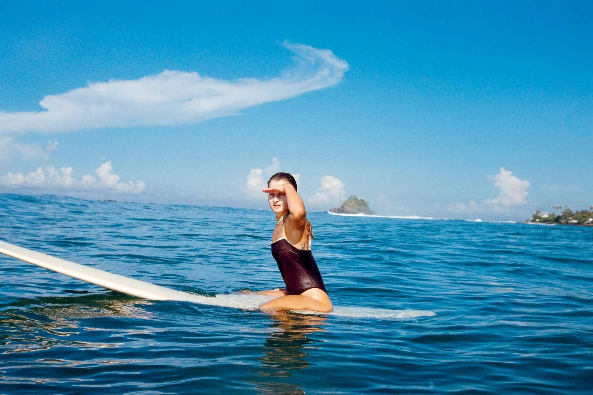 A woman in a black swimsuit sitting on a paddleboard in the ocean coastline with an island and partly cloudy sky in the background