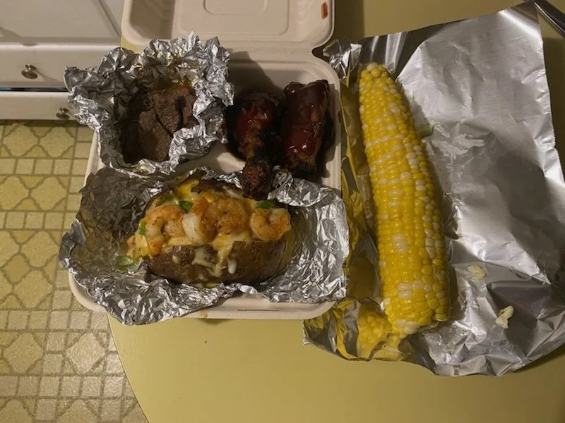 Baked potato topped with cheese, shrimp, and other toppings, three barbecue chicken drumsticks, and an ear of corn on the cob, all in aluminum foil, on a yellow table.