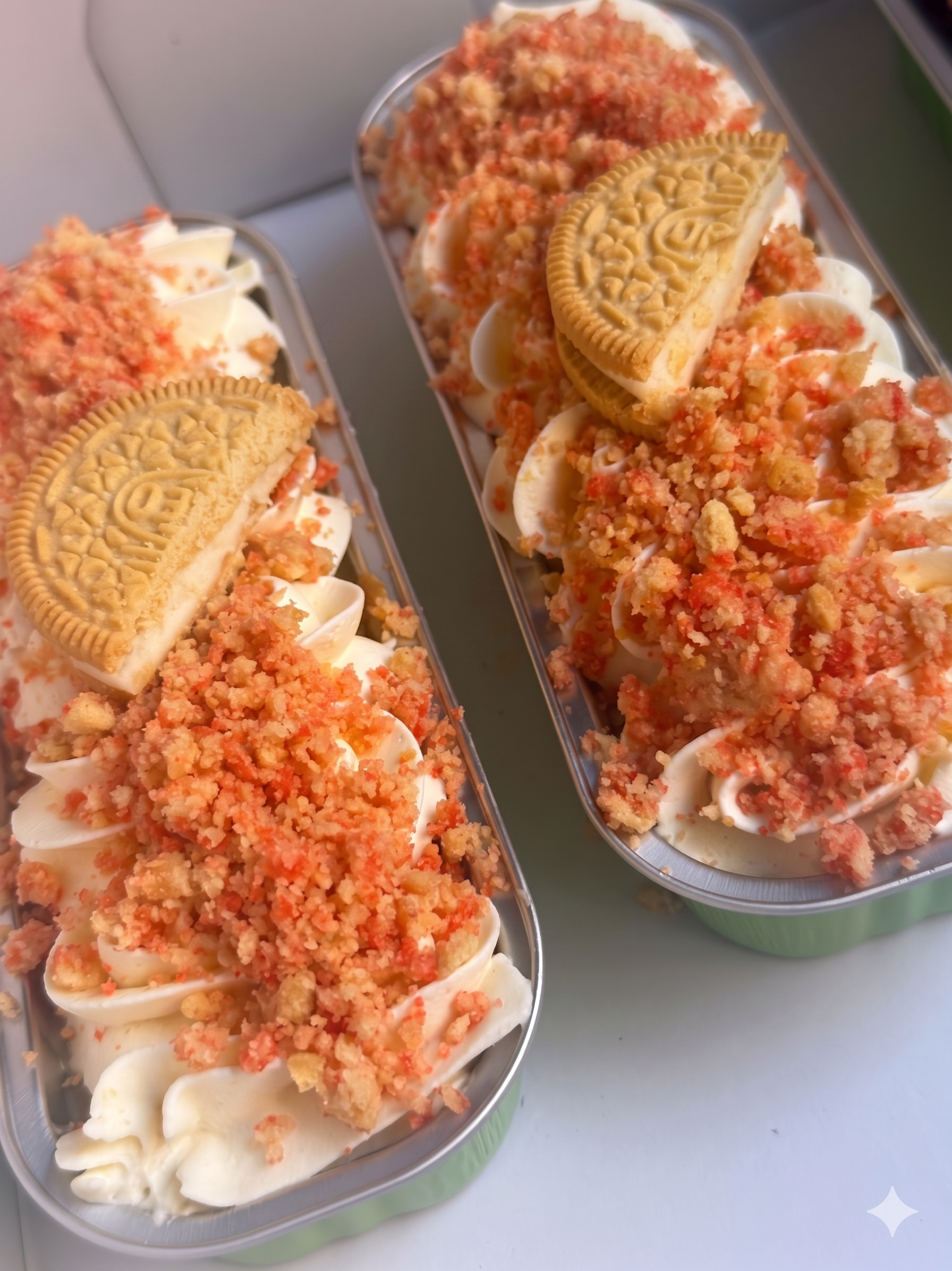 Two clear plastic containers filled with vanilla ice cream, topped with crushed cookies and crumble, placed on a white surface.