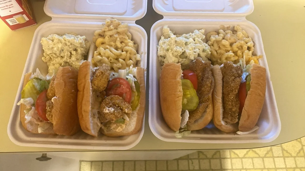 Two foam containers filled with fried fish sandwiches, macaroni and cheese, and potato salad, on a light yellow table.