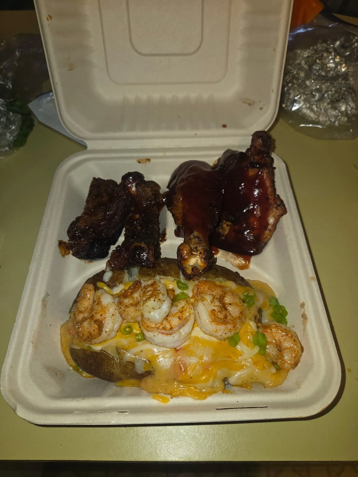 Takeout container with barbecued ribs, shrimp and cheese stuffed potato, and probably baked beans or other side dishes.