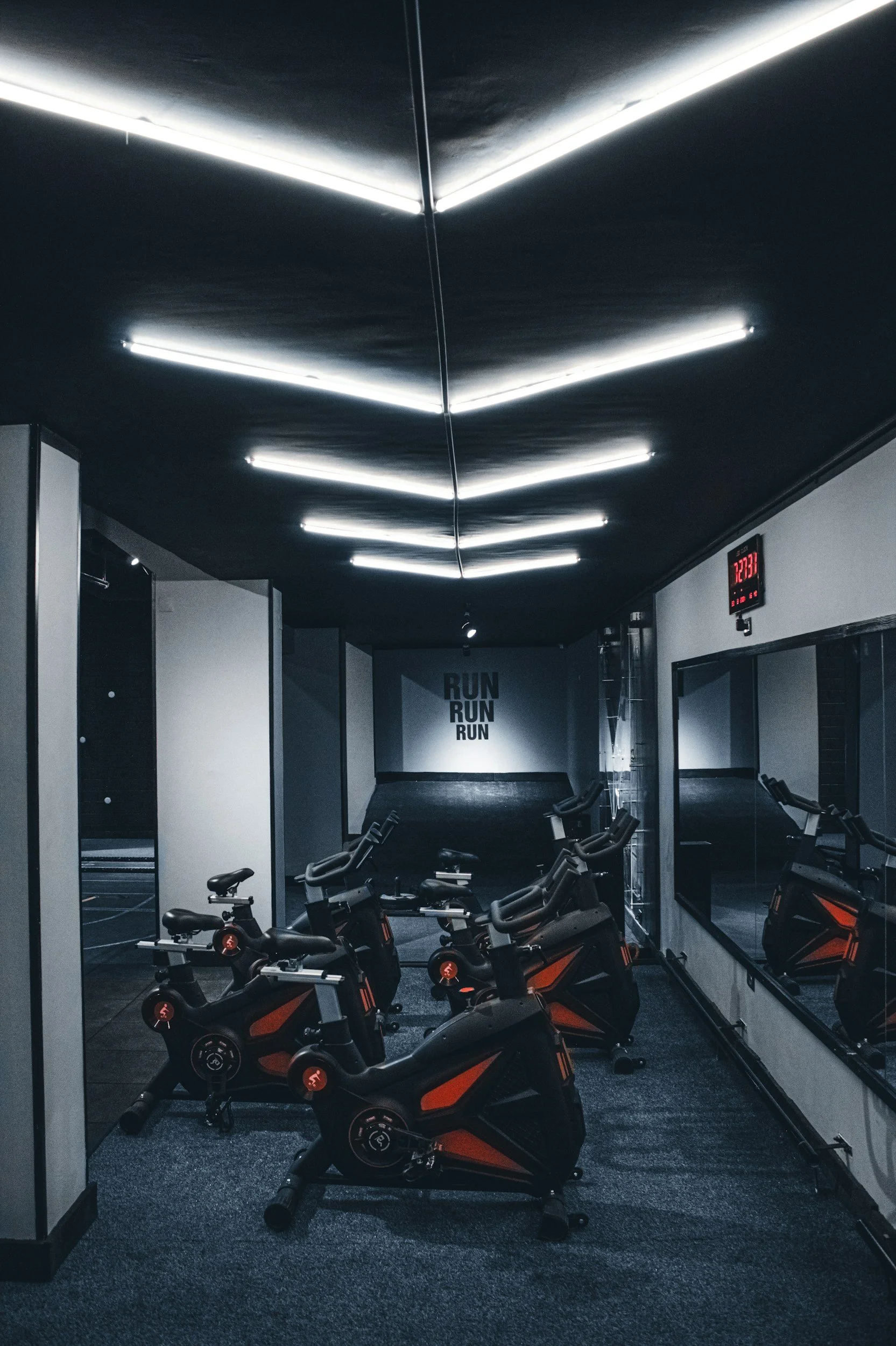 Salle de sport avec des vélos d'appartement alignés devant un miroir. Mur avec la phrase 'RUN RUN RUN' et un affichage numérique lumineux indiquant 12:33.