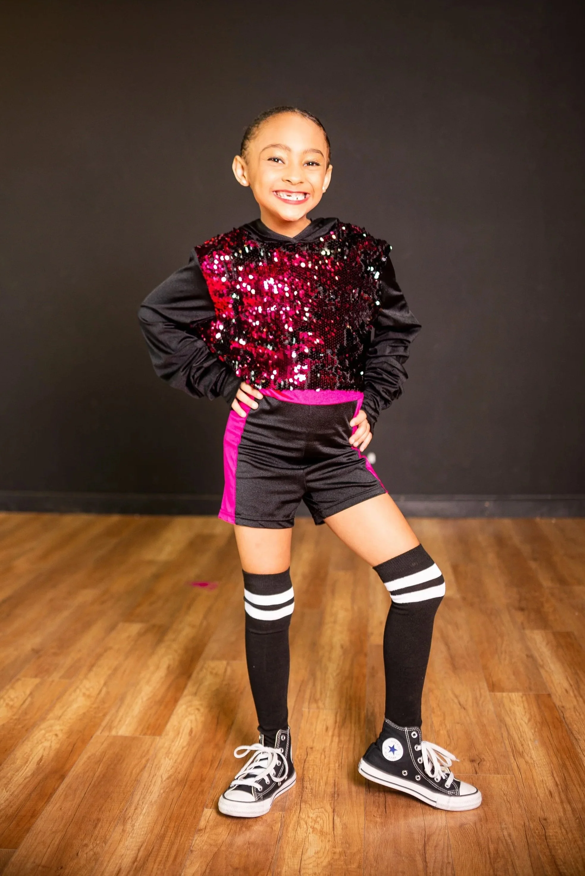 A young girl with a big smile, standing indoors on a wooden floor against a dark background. She is wearing a black and pink athletic outfit, high-top sneakers, and striped knee-high socks.