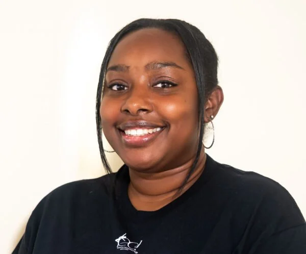 A woman with dark hair, wearing a black shirt, smiling at the camera against a plain, light-colored background.