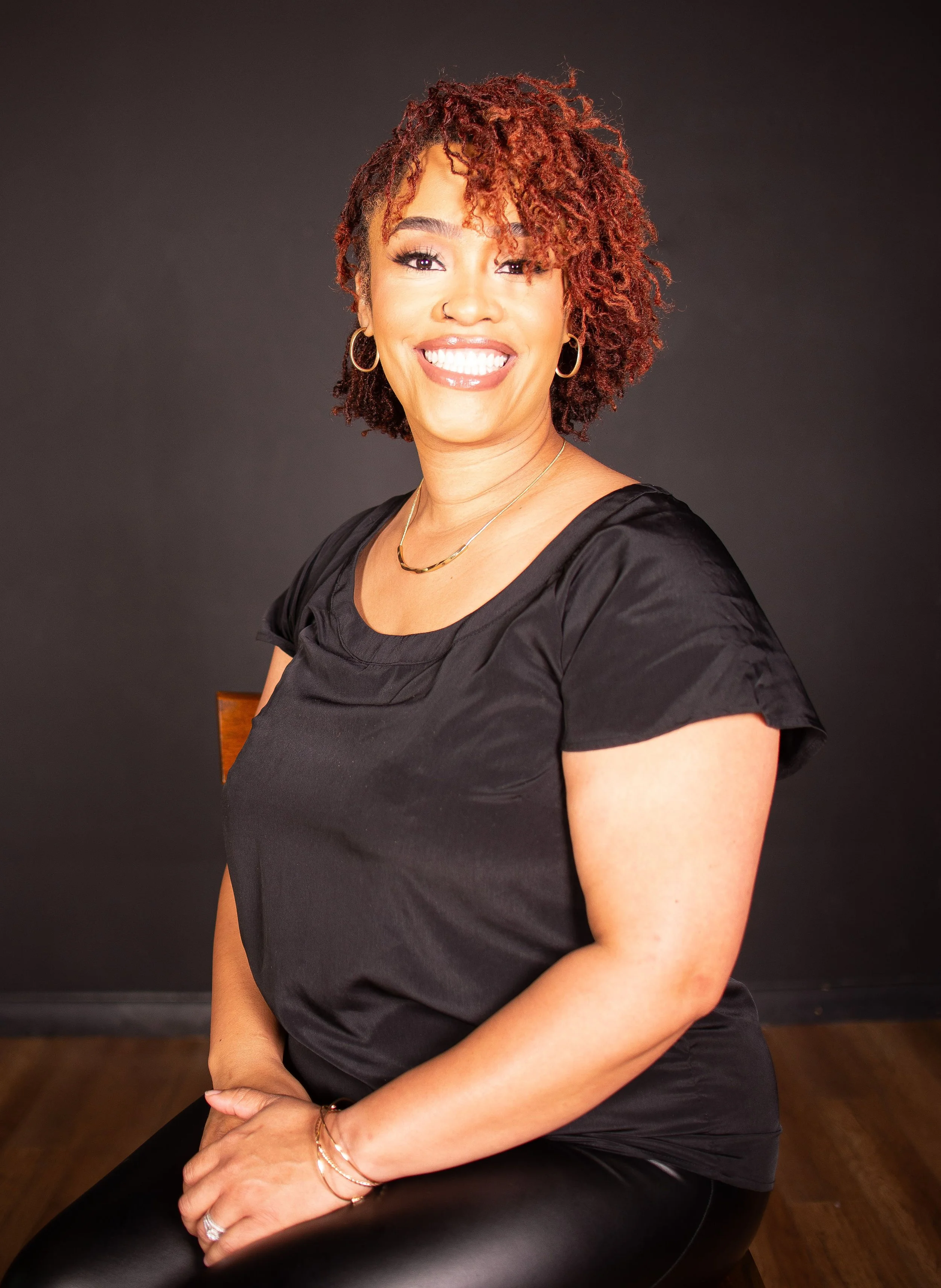 A woman with curly, reddish-brown hair smiling and wearing a black top, silver hoop earrings, and a gold necklace, seated against a black background.