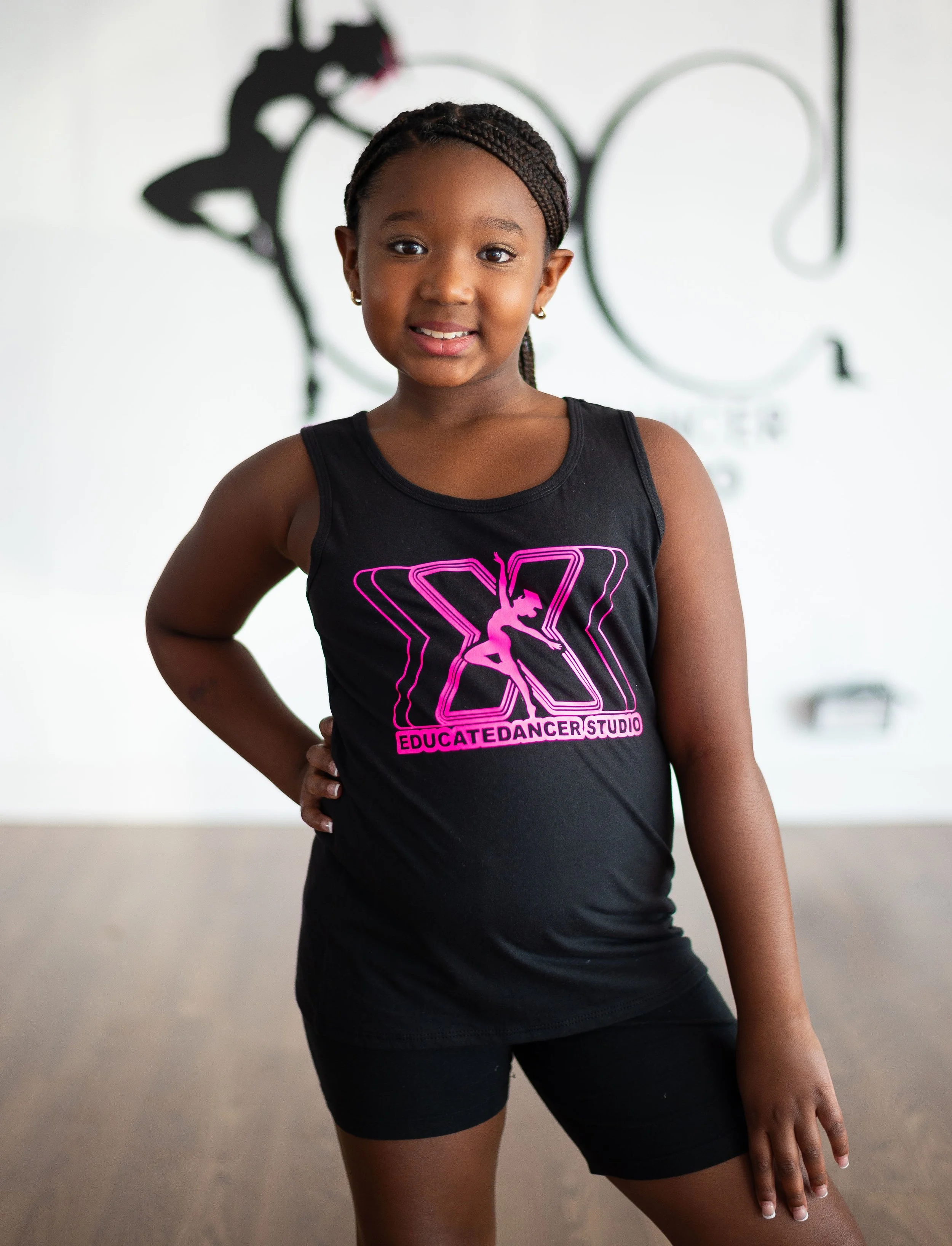 Young girl standing in a dance studio, wearing a black tank top with pink logo and text, smiling at the camera.
