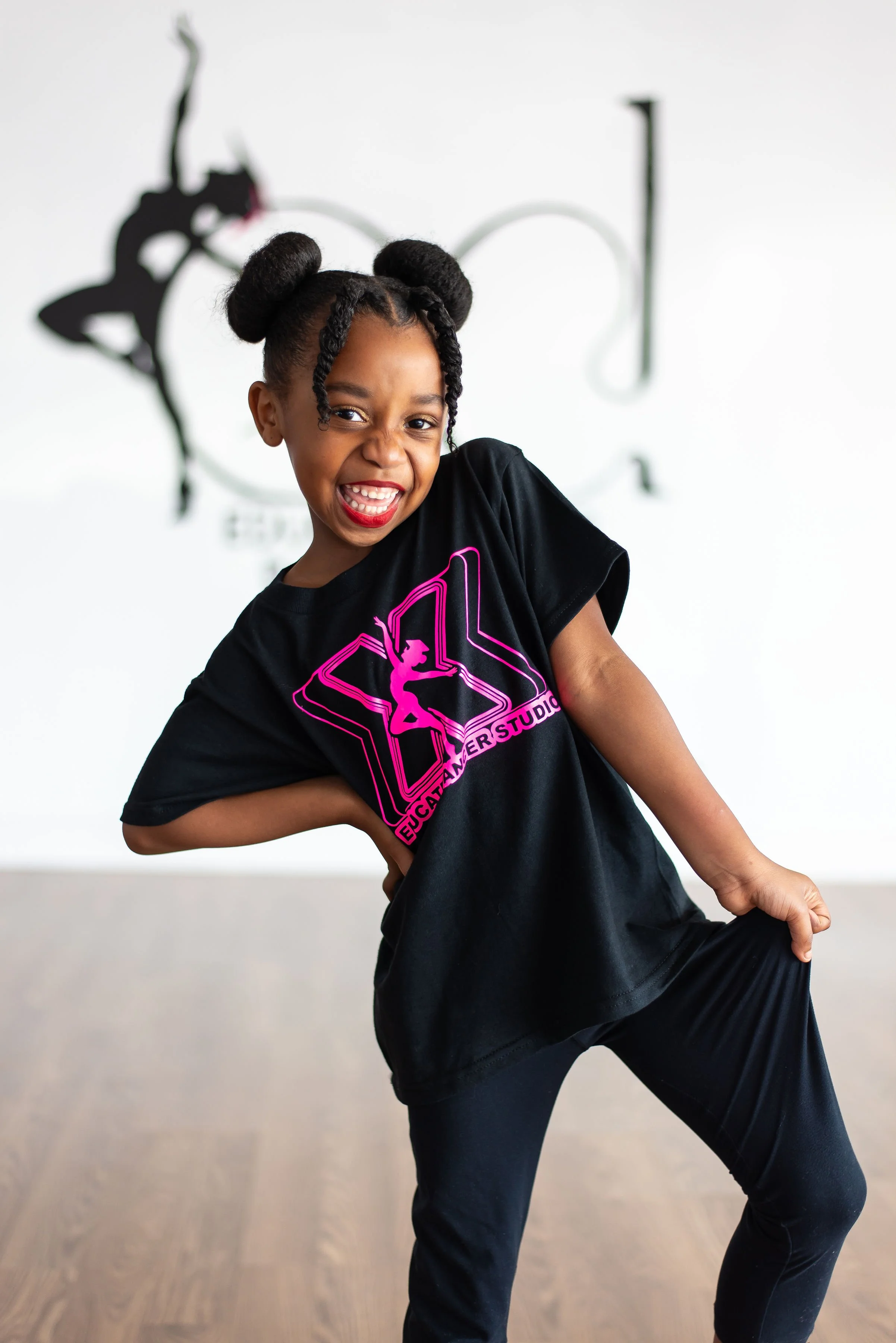 A young girl with braided hair styled in two buns is smiling and posing in a dance studio. She is wearing a black T-shirt with a pink graphic and text that reads 'EDUCATOR STUDIO' and black pants. The background is white with a black and white wall decoration of a dancer silhouette.