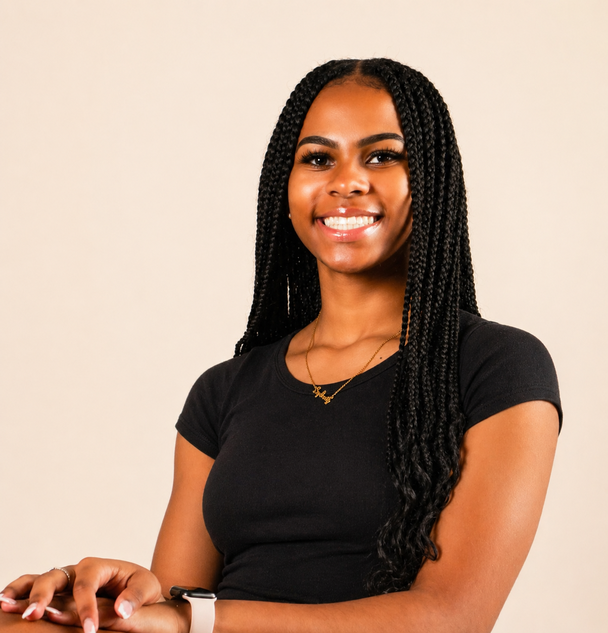 A young woman with long braided hair smiling, wearing a black t-shirt, a gold necklace, and a white wristwatch, against a plain light-colored background.