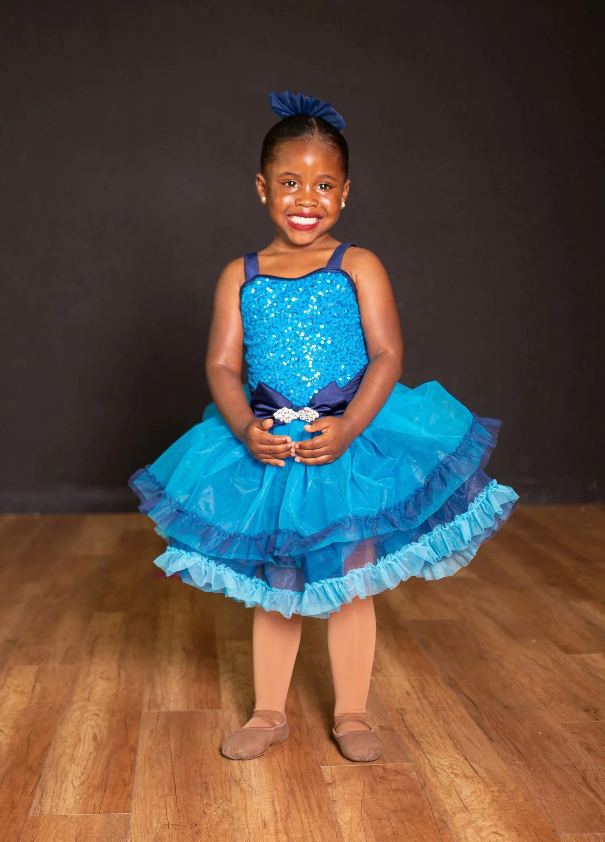 A young girl dressed in a vibrant blue tutu dance costume with a matching bow in her hair, smiling confidently in front of a black background.