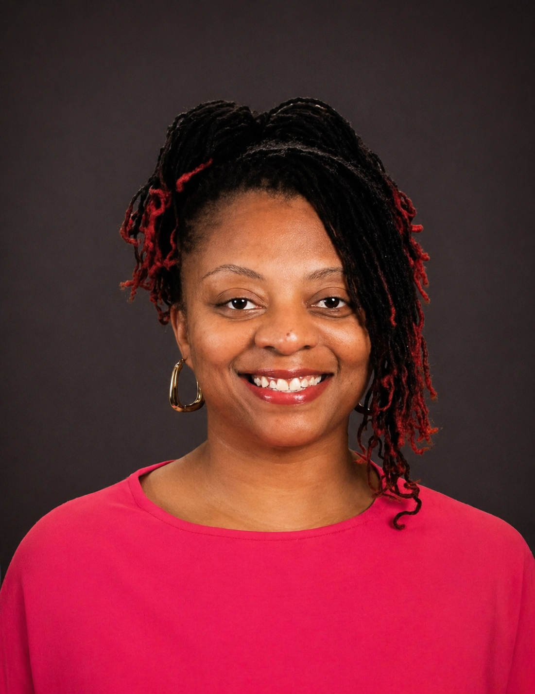 A smiling woman with dark curly hair with red highlights, wearing a pink top and gold hoop earrings, standing against a dark background.