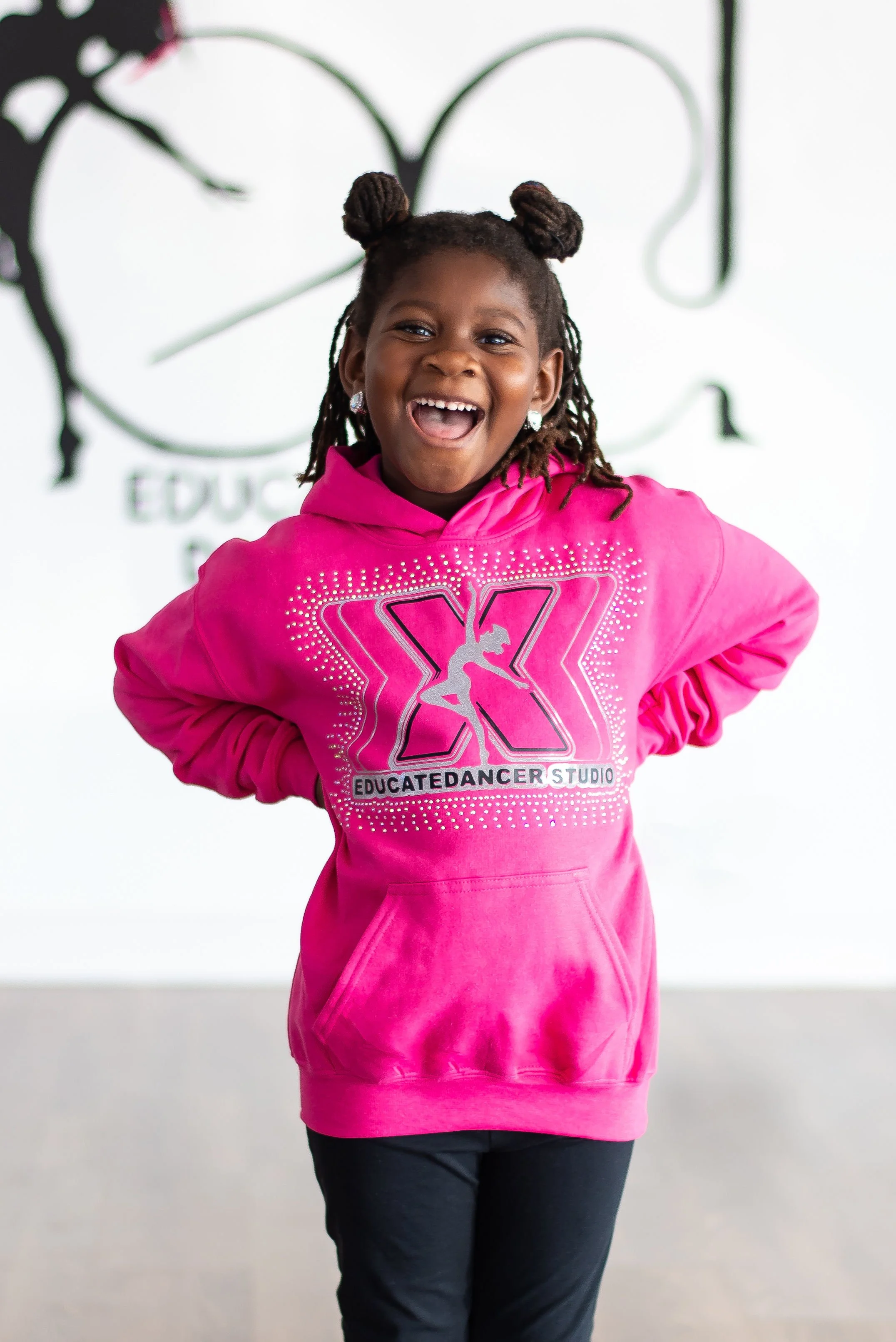 A young girl with dark skin and styled dreadlocks wearing a bright pink hoodie with the logo 'X' and the words 'Educated Dancer Studio' stands in front of a white wall with part of a logo or design visible in the background. She is smiling and has her hands on her hips.