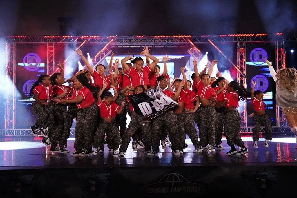Group of young girls performing a dance on stage during a competition, wearing matching red shirts and camouflage pants, with stage lights and backdrop in the background.