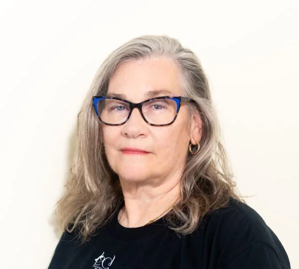 A mature woman with gray hair, wearing glasses with black and blue frames, earrings, and a black shirt, standing against a plain light background.