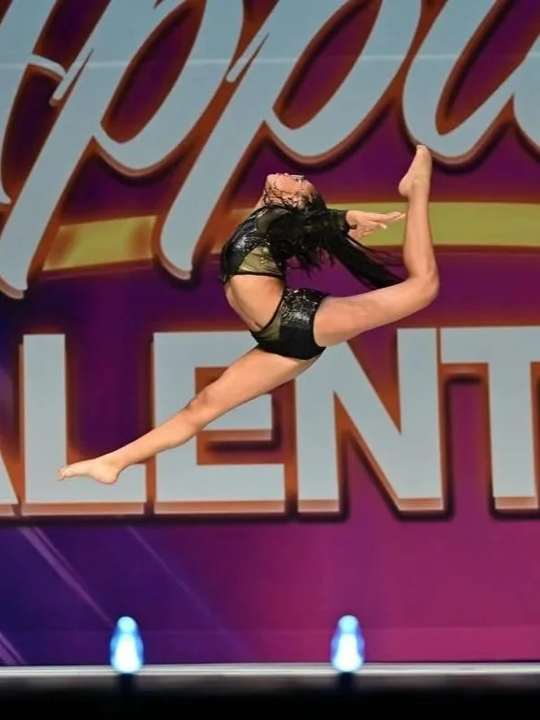 A female gymnast performing a leap with a split position during a dance or fitness competition on stage.