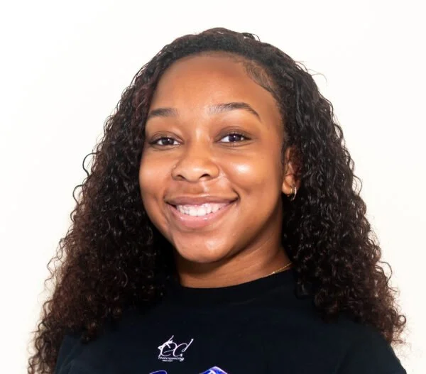 Smiling young woman with long curly hair and small hoop earrings, wearing a black t-shirt against a white background.